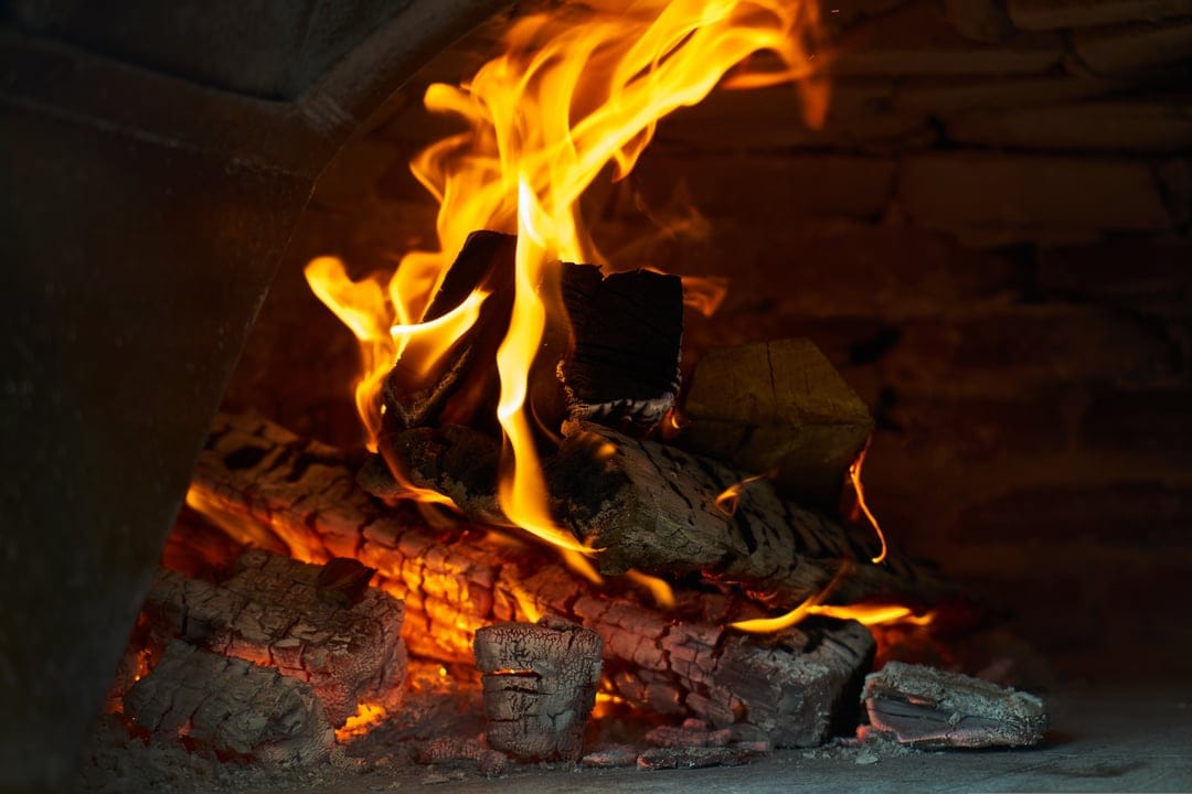 Roaring flames and glowing embers flicker between charred logs inside a rustic stone-lined fireplace interior.