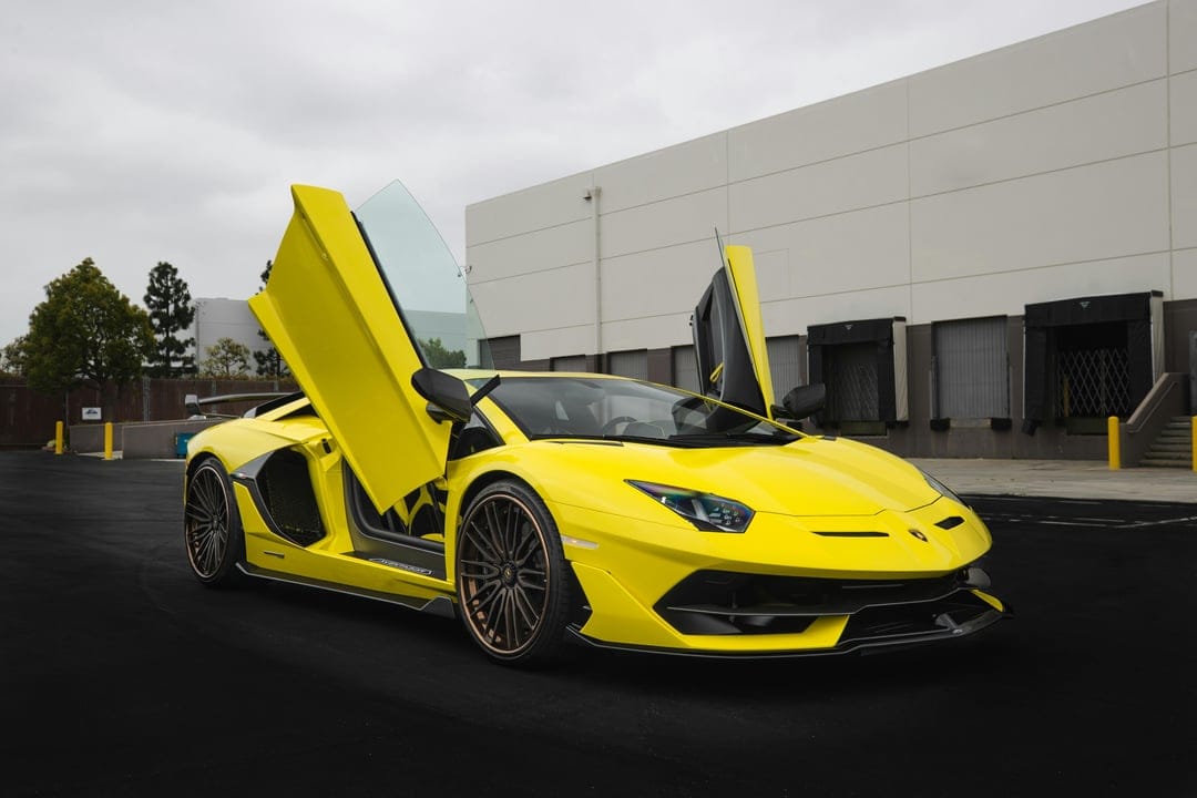 Yellow Lamborghini Aventador with upward-opening scissor doors parked on asphalt outside an industrial building.