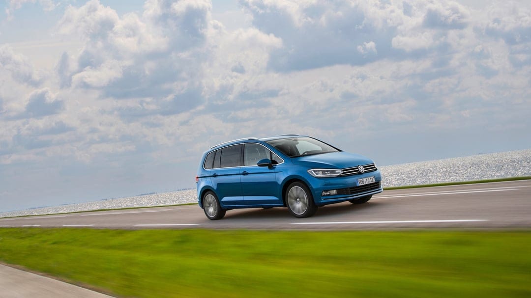 Blue Volkswagen Touran driving on a highway by the sea under a bright, cloud-filled sky with motion blur.