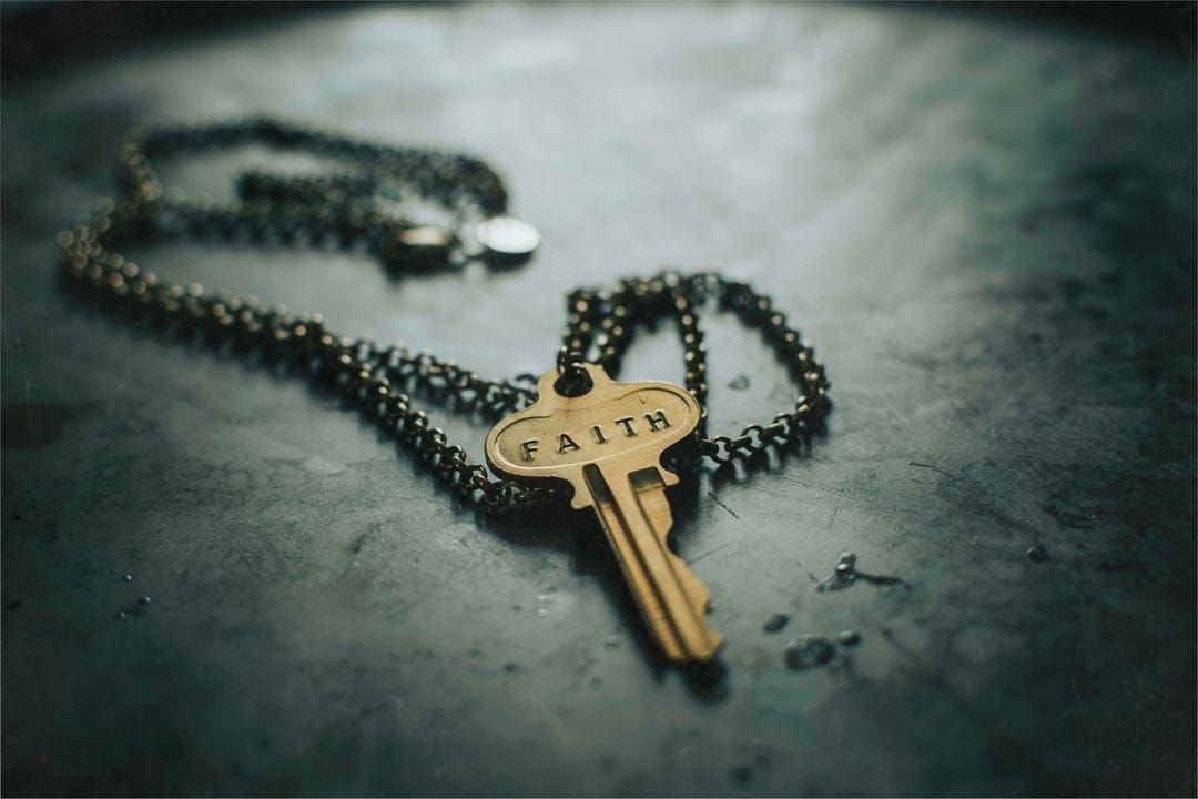Golden key pendant with faith engraving on a dark textured metallic surface with a blurred dark chain backdrop.
