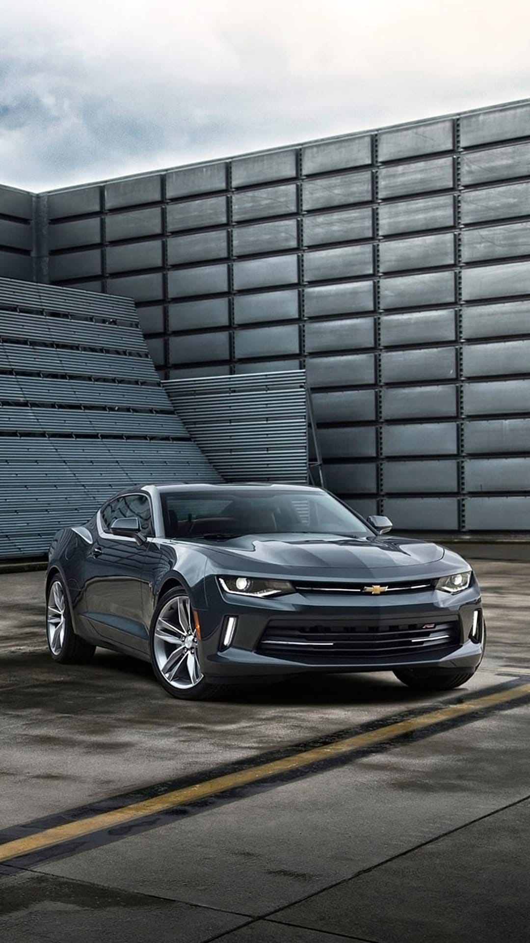 Dark grey Chevrolet Camaro parked on wet asphalt in front of a modern industrial building with textured walls.