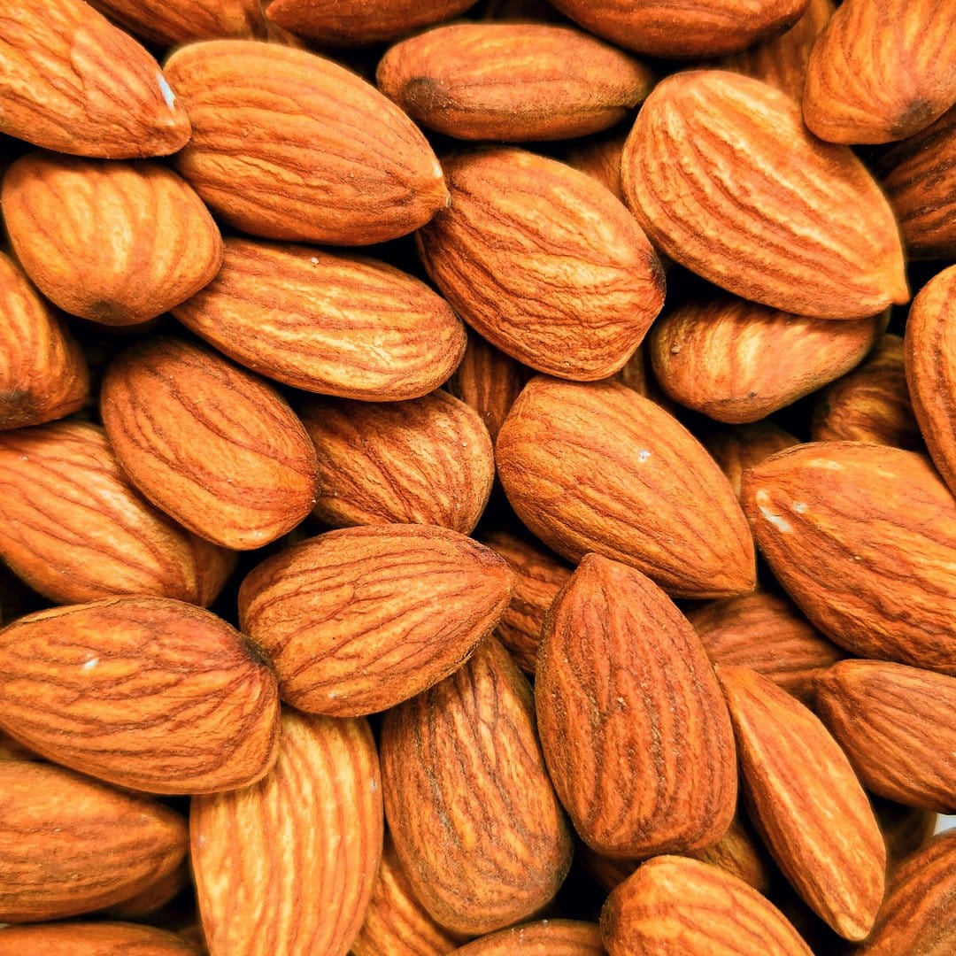 Raw almonds with textured brown skins are piled together in a high-angle macro shot showing natural ridges.