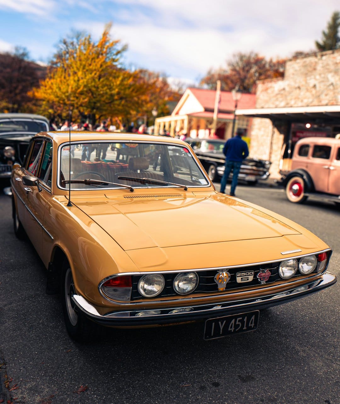 Golden Triumph Stag classic car with chrome details parked on a street lined with colorful autumn leaves.