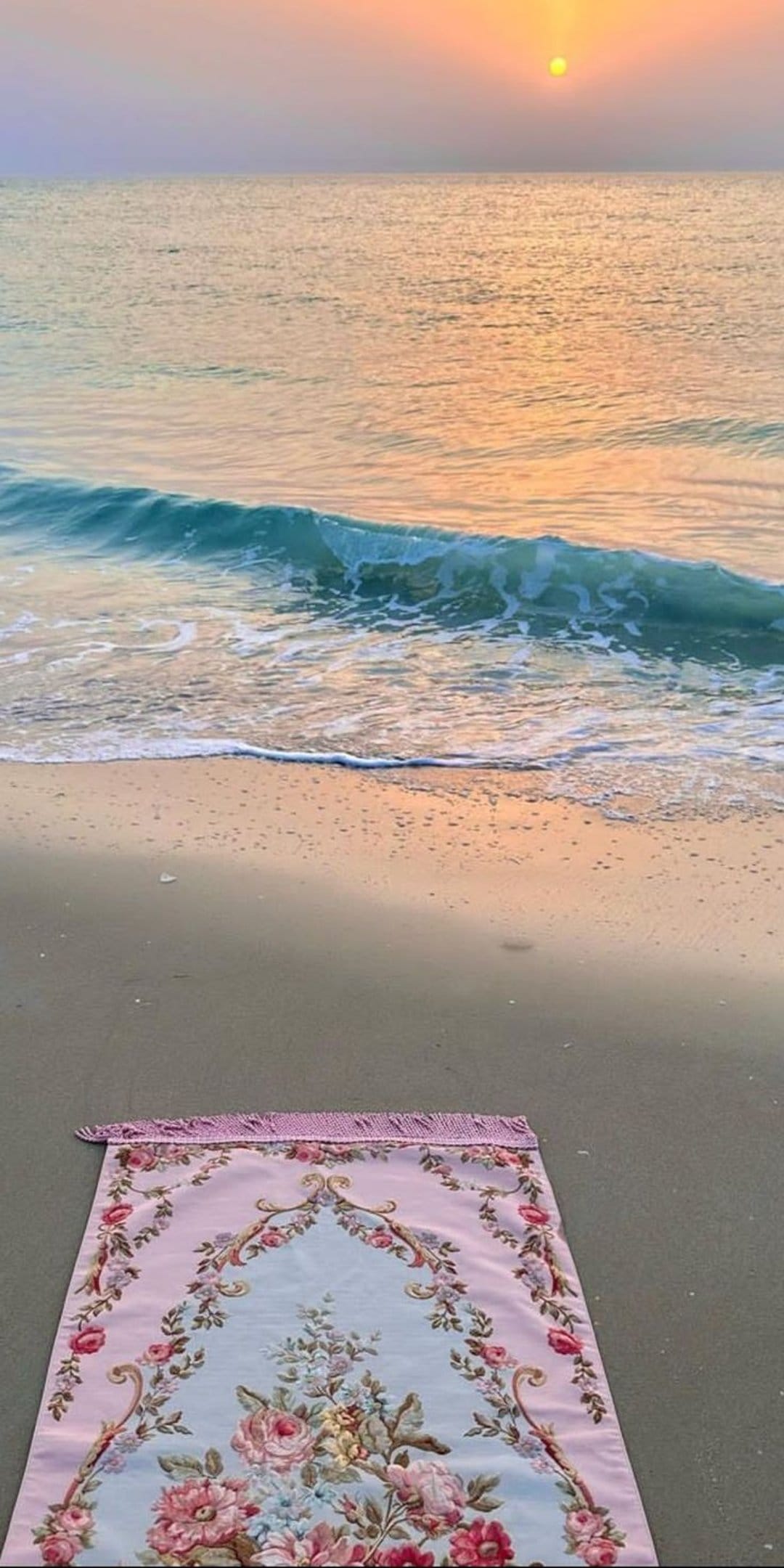 Woven prayer rug lies on wet sand as the sun sets over ocean waves under a vibrant orange and purple sky.