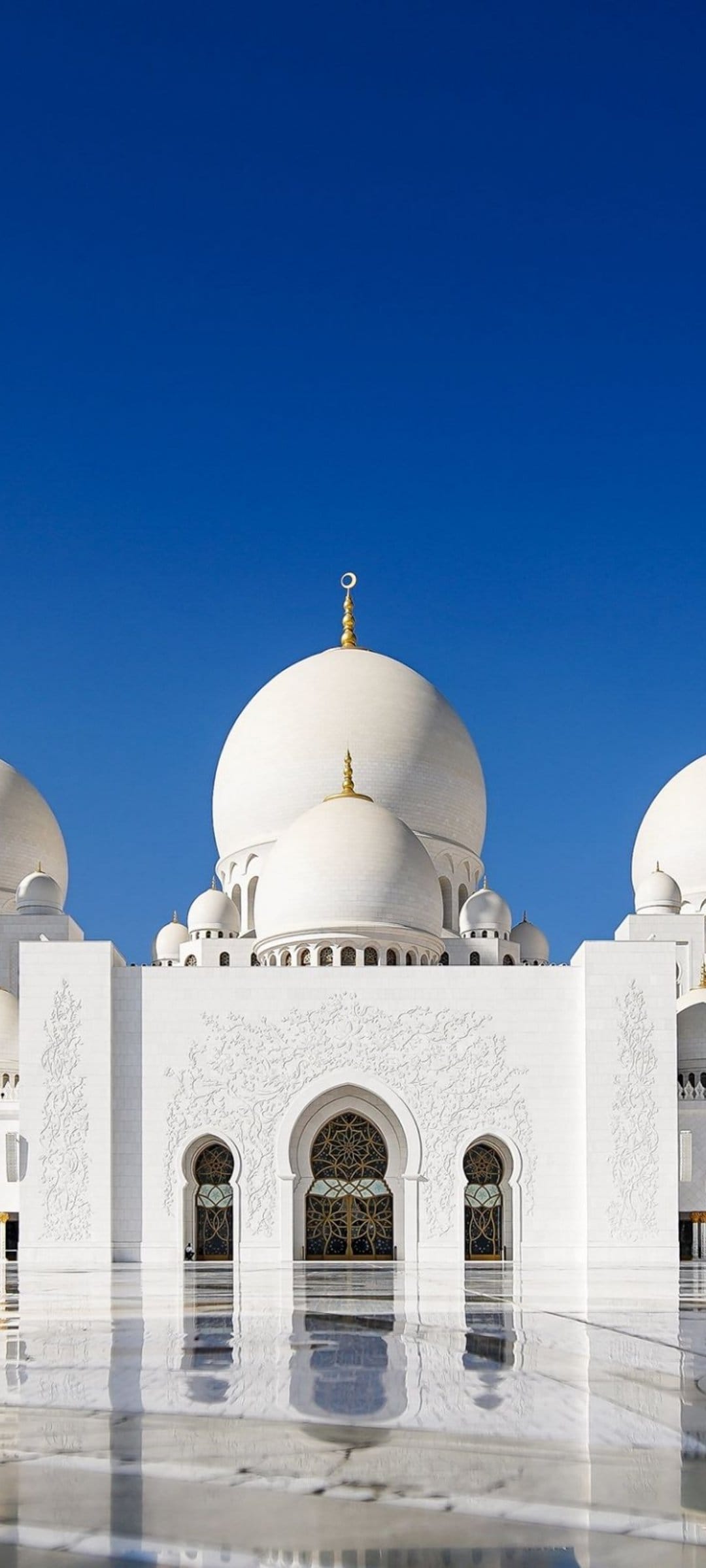 Sheikh Zayed Grand Mosque white domes reflect on a wet marble floor under a clear blue sky in Abu Dhabi.