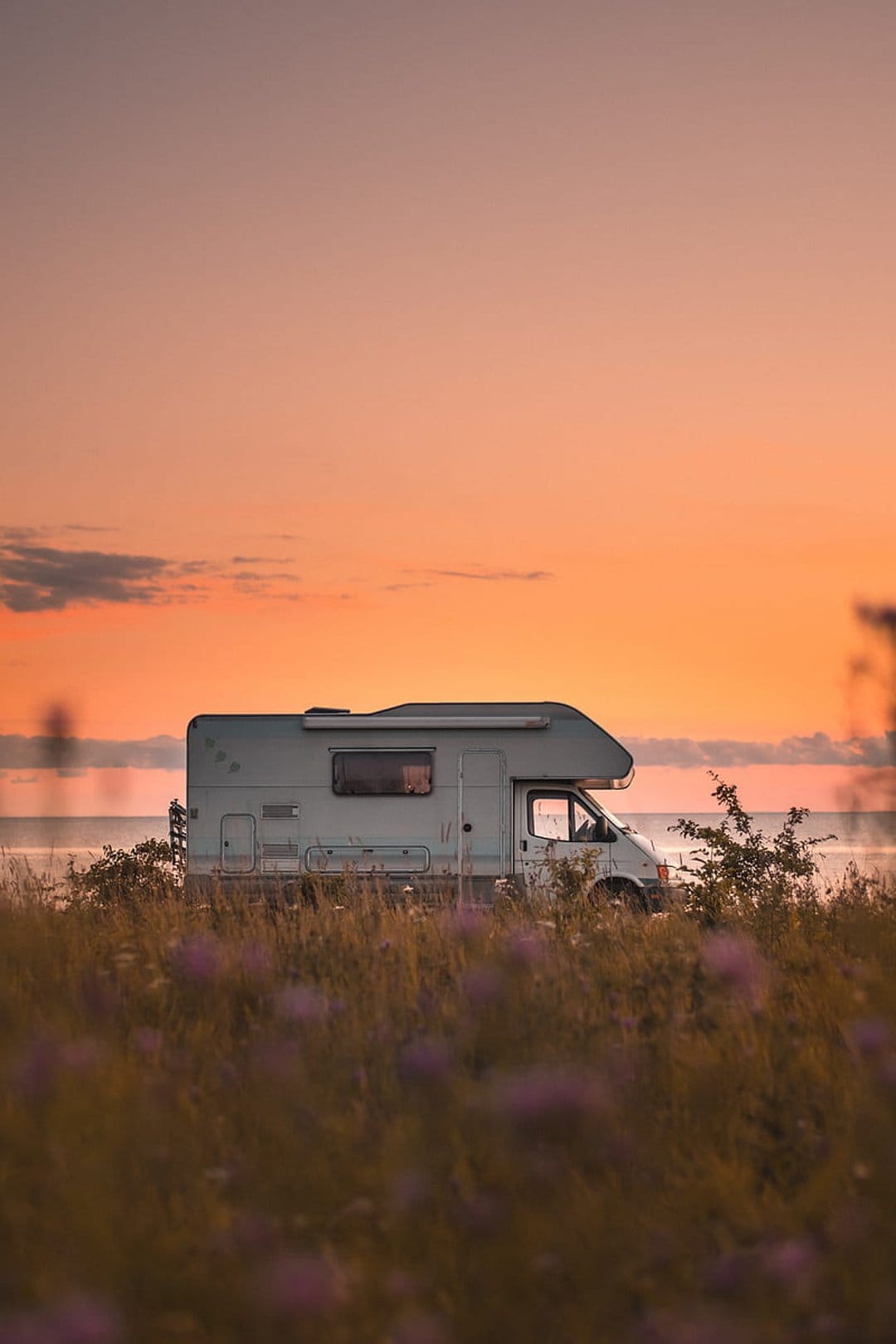 White RV camper van parked in tall grass overlooking a coastal sunset with vibrant orange and pink clouds.