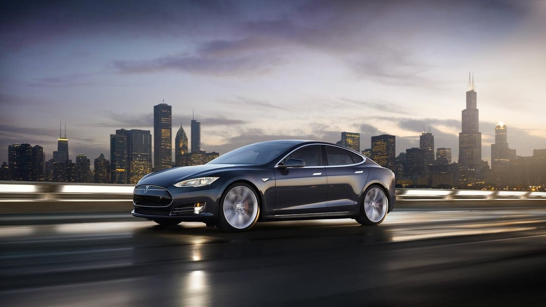 Tesla Model S speeding on a wet city road at dusk with the Chicago skyline glowing in the background.