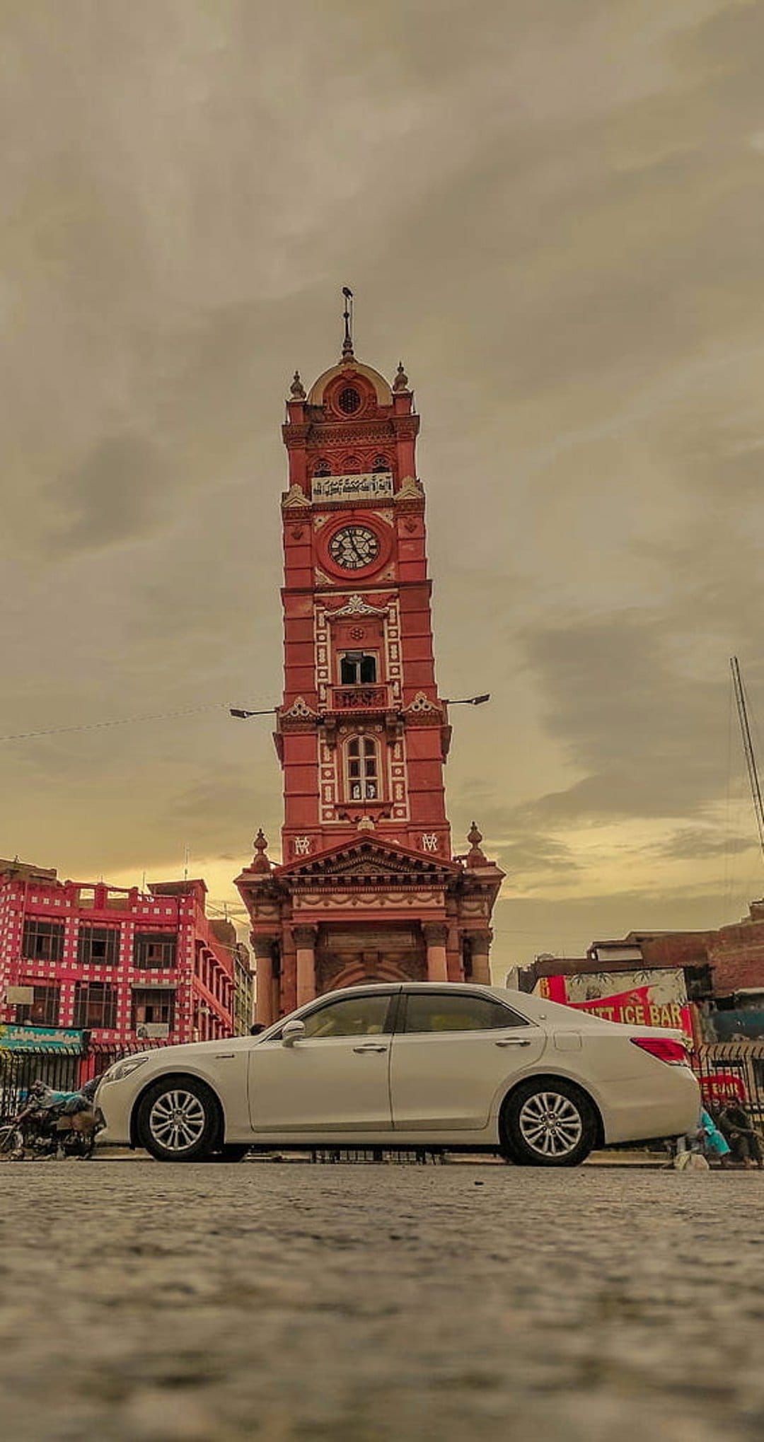White sedan parked in a city square beneath a tall red brick clock tower and a hazy overcast sky.