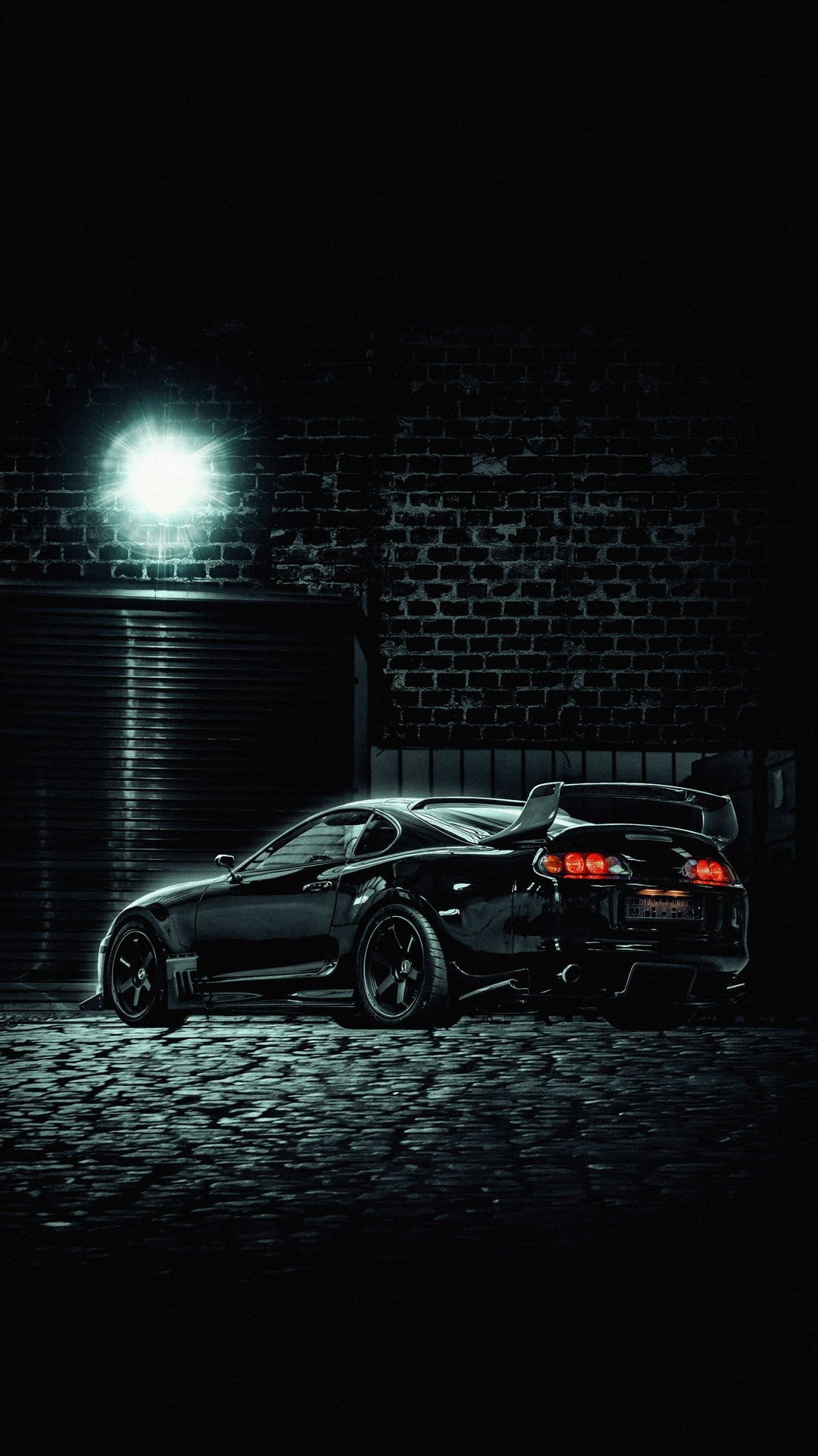 Black Toyota Supra parked on wet cobblestones in a dark alley illuminated by cool blue neon light from a brick wall.