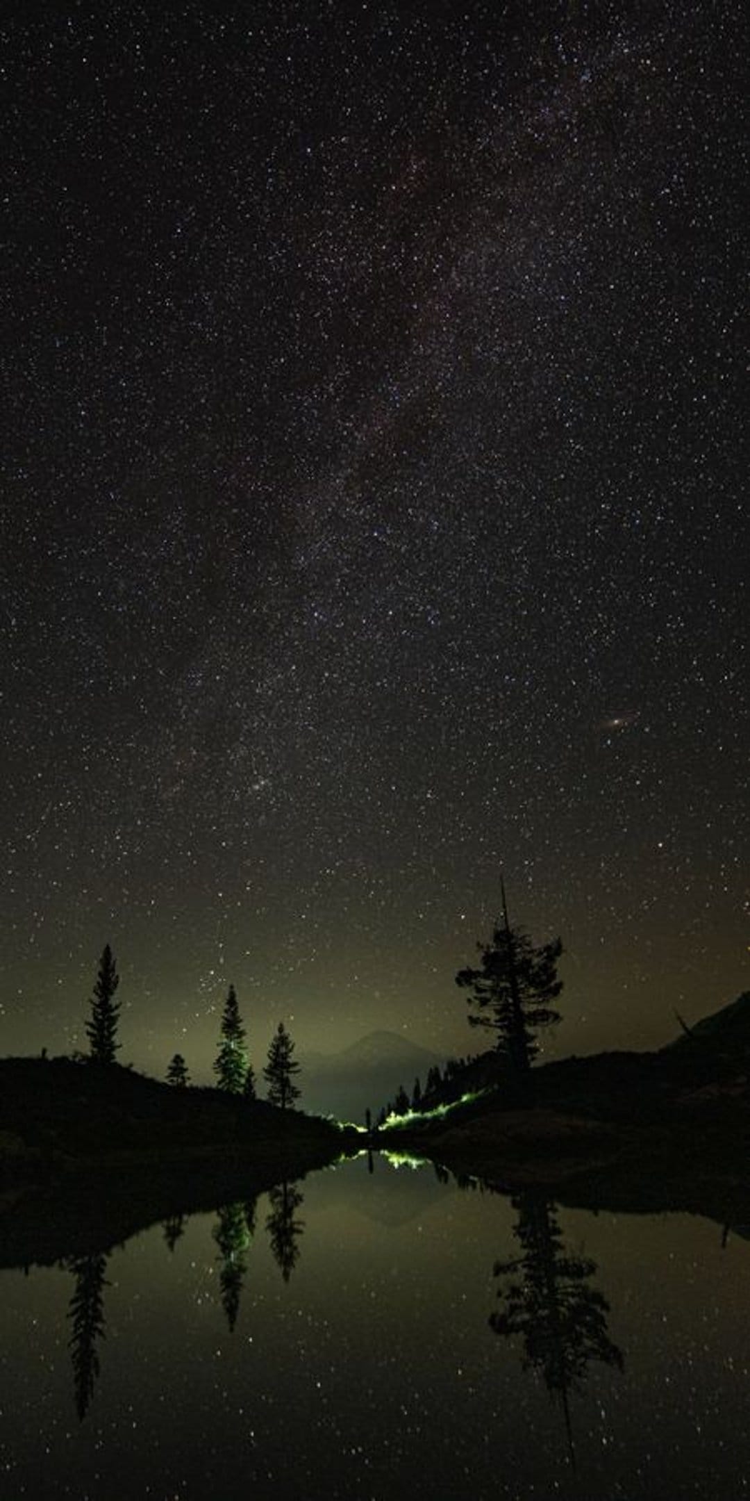 Milky Way galaxy core reflected in a still lake at night with evergreen tree silhouettes on the shore.