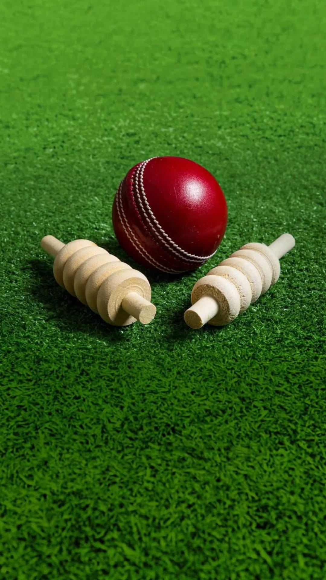 Red cricket ball and two wooden bails rest on a vibrant green grass pitch in a sharp macro composition.