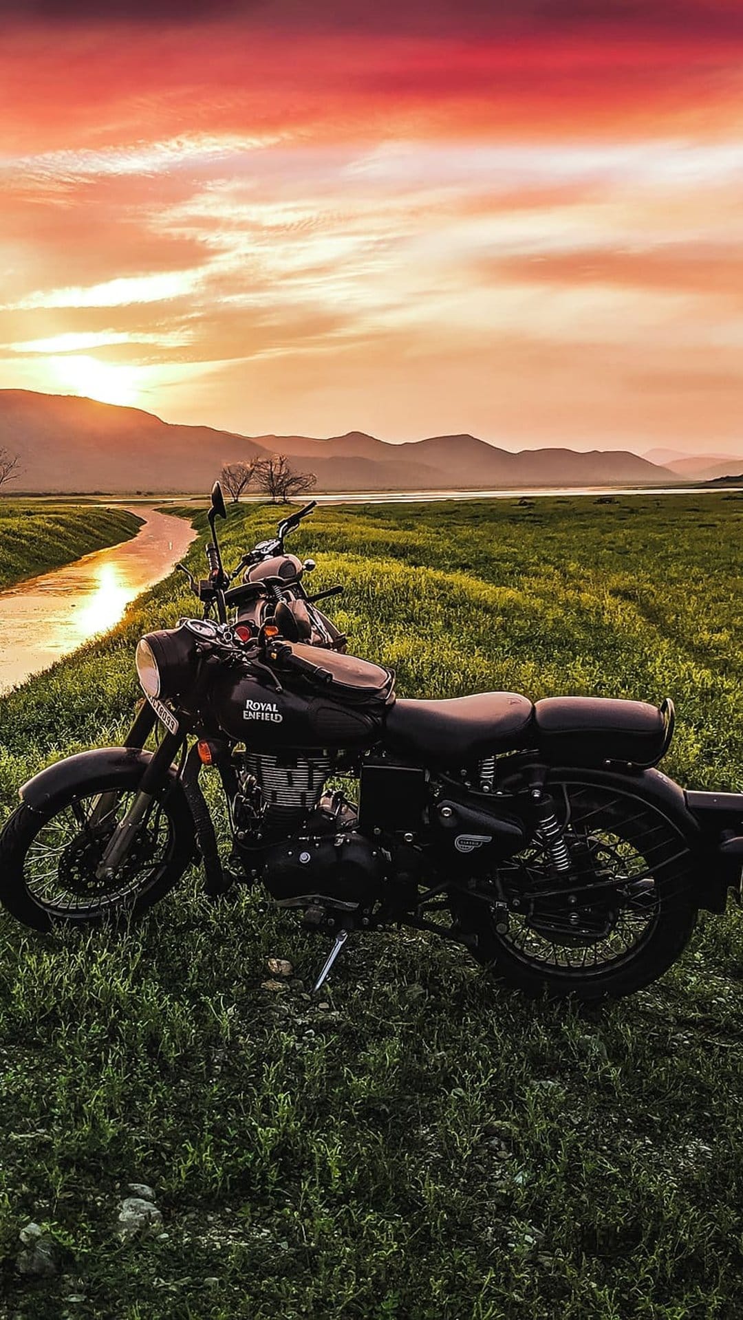 Black Royal Enfield bike stands near a winding creek under a fiery sunset sky with distant peaks in the background.