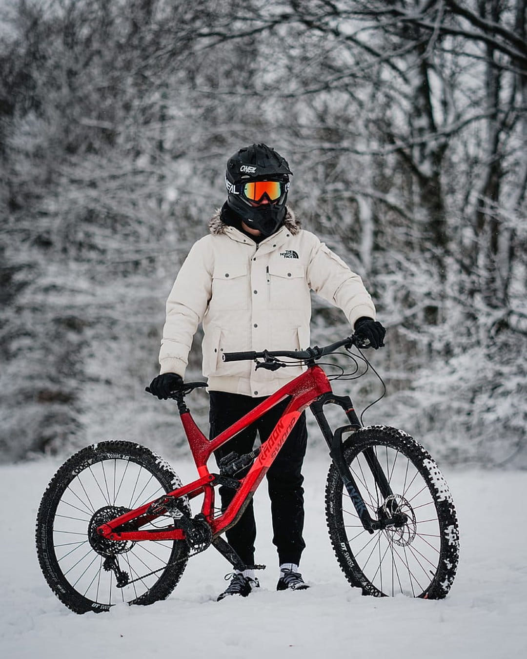 Mountain biker in a white jacket holds a red bike on a snowy trail surrounded by frost-covered evergreen trees.