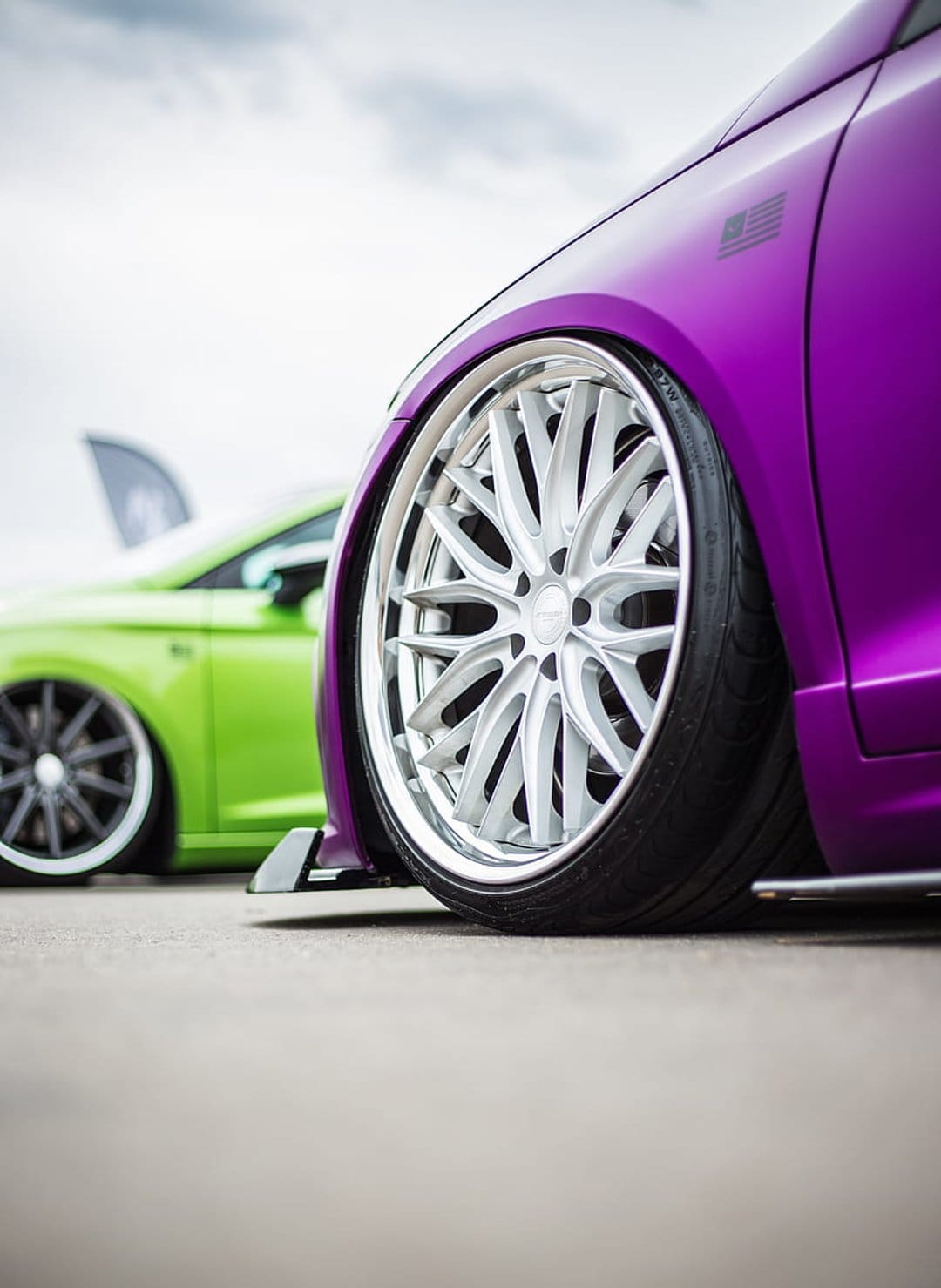 Silver alloy lowrider rims on purple and green customized cars show detailed chrome spokes and a lowered stance.