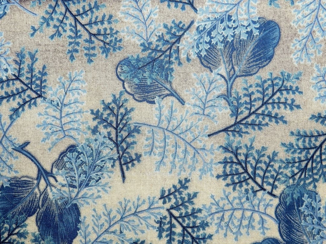Blue fern fronds and stylized leaves overlap on a textured beige fabric surface with a natural fiber grain.