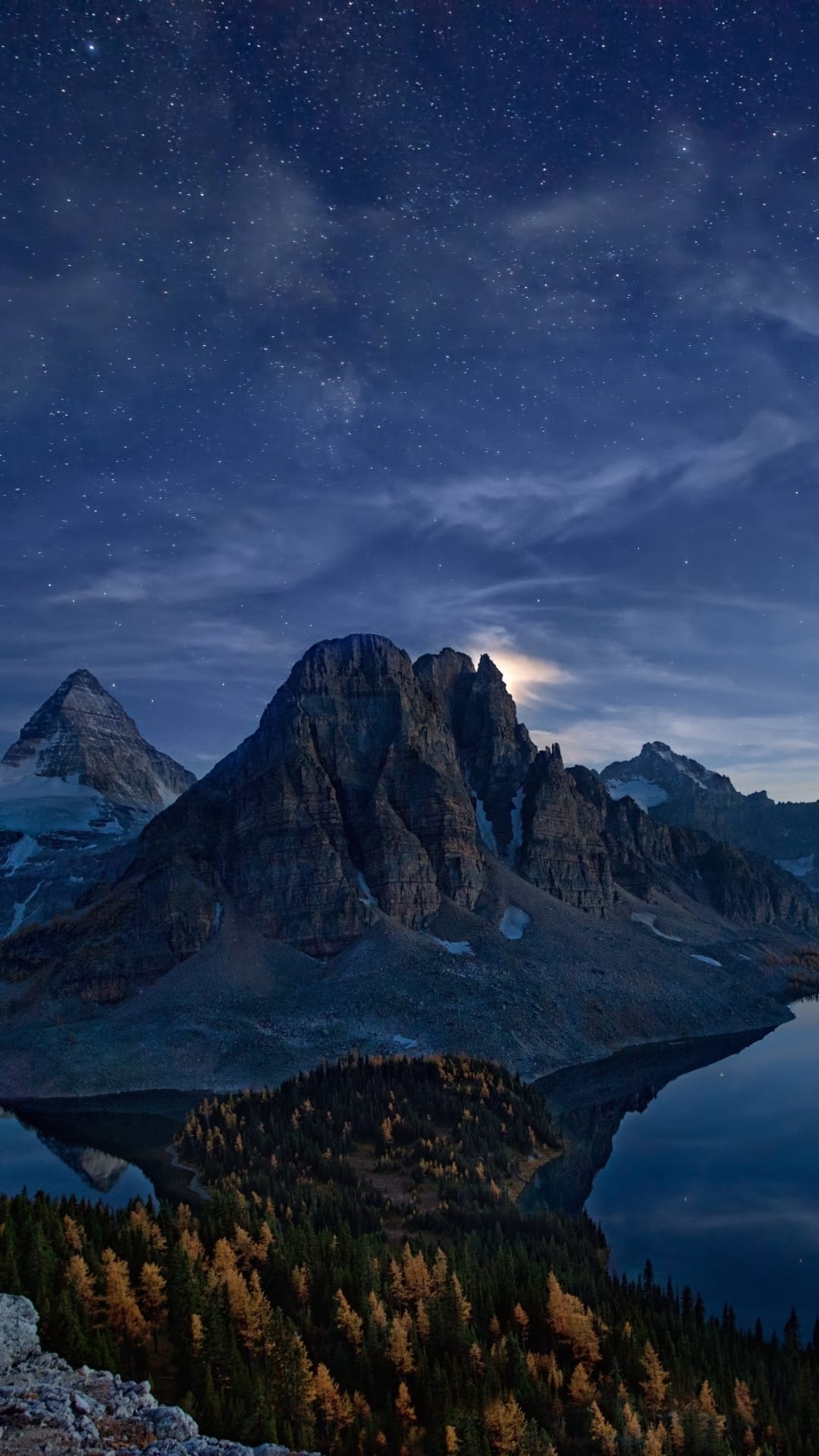 Snow-capped mountain peaks reflect in a still lake under a starry night sky with orange autumn trees on the shore.