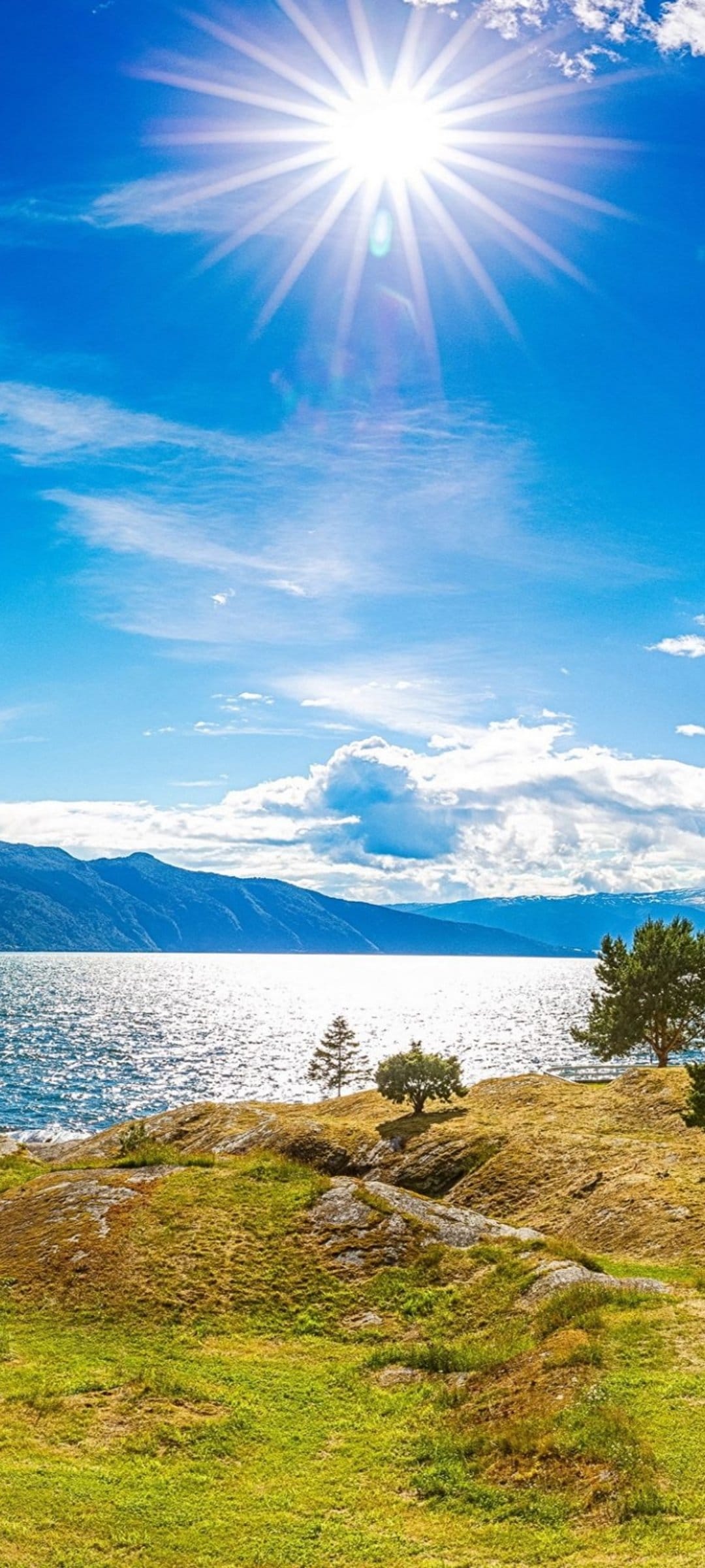Distant mountain peaks rise above a sparkling lake and grassy hills beneath a brilliant sun in a clear sky.