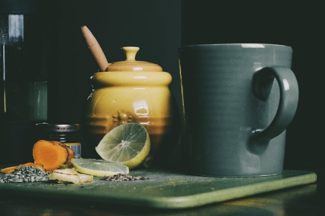Turmeric root, ginger slices, and a lemon half sit beside a honey dipper on a dark green board.