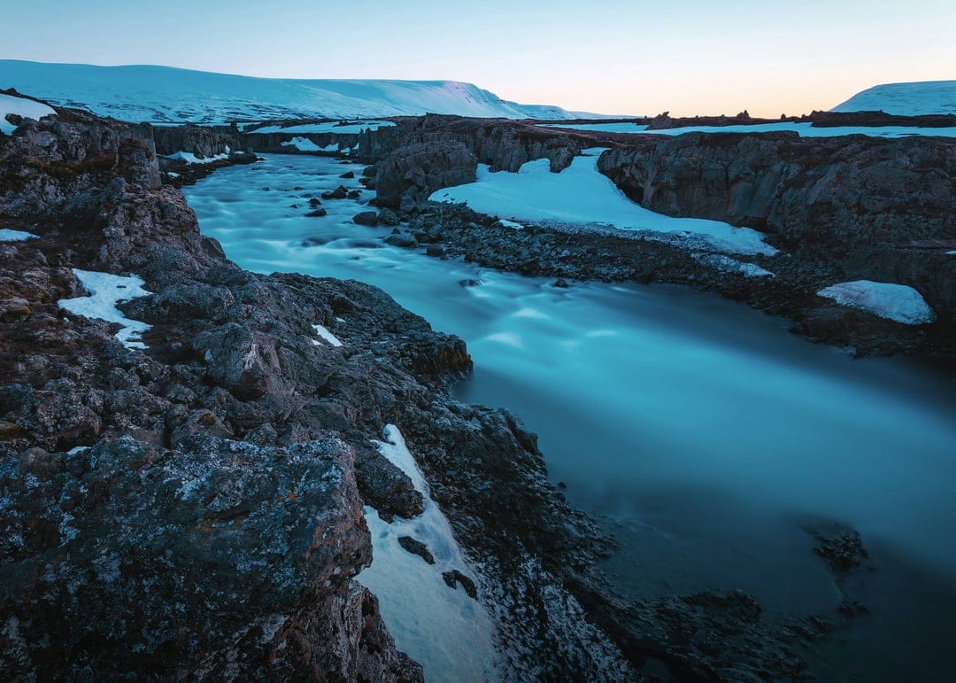 Blue river water flows between snow-covered volcanic rocks under soft light in a vertical Icelandic landscape.