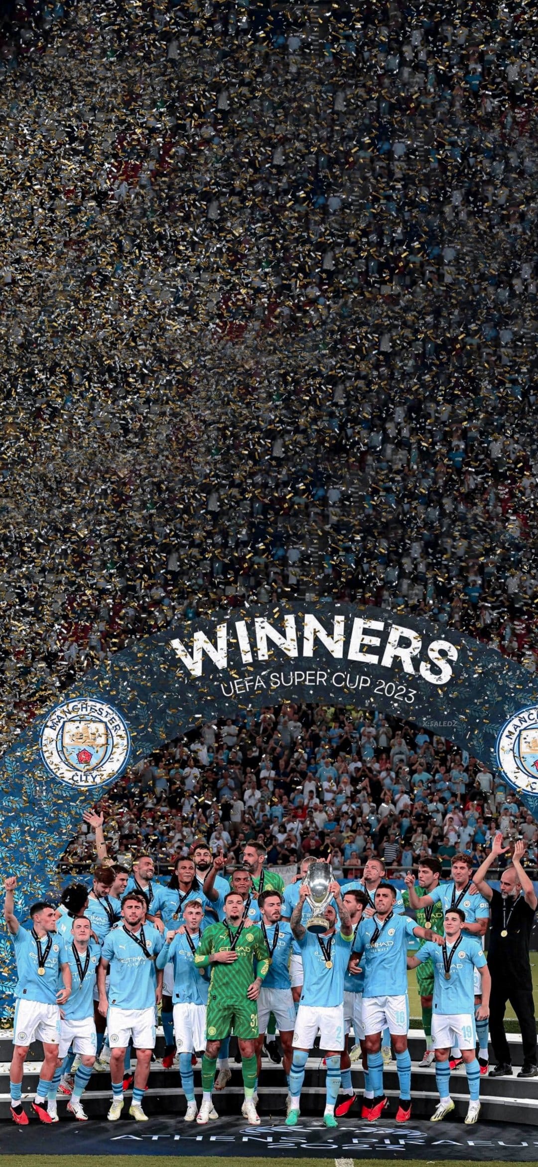 Manchester City football players raise a silver trophy during a night celebration with blue and white confetti.