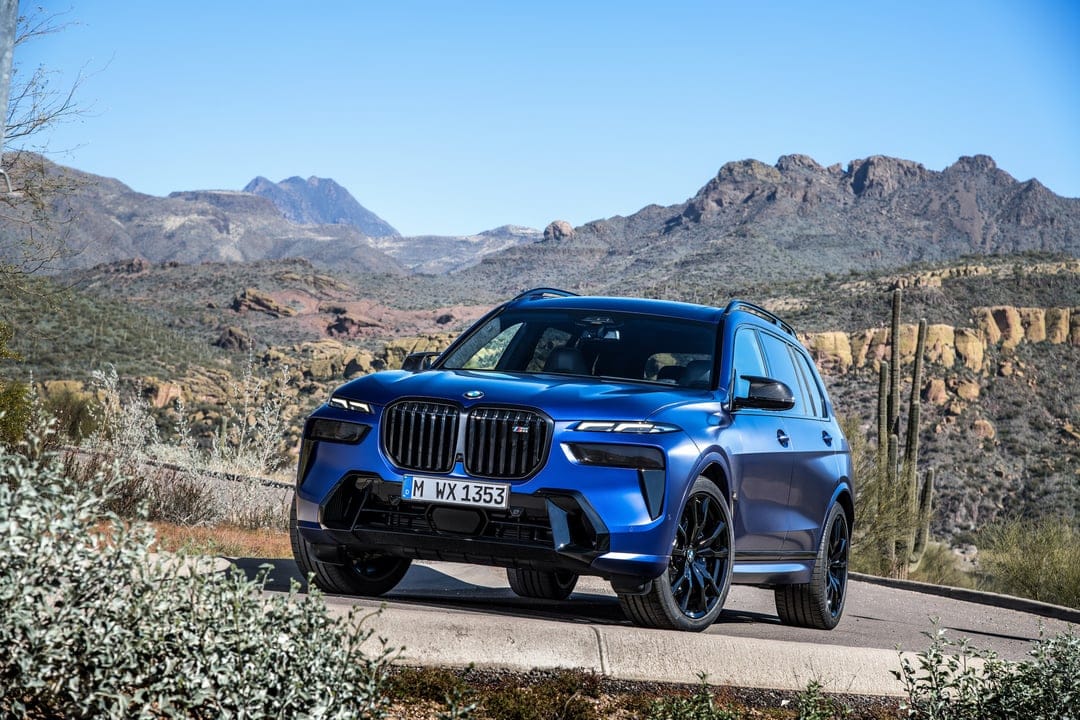 Blue BMW X7 luxury SUV on a desert mountain road with rocky peaks and saguaro cacti in the background.