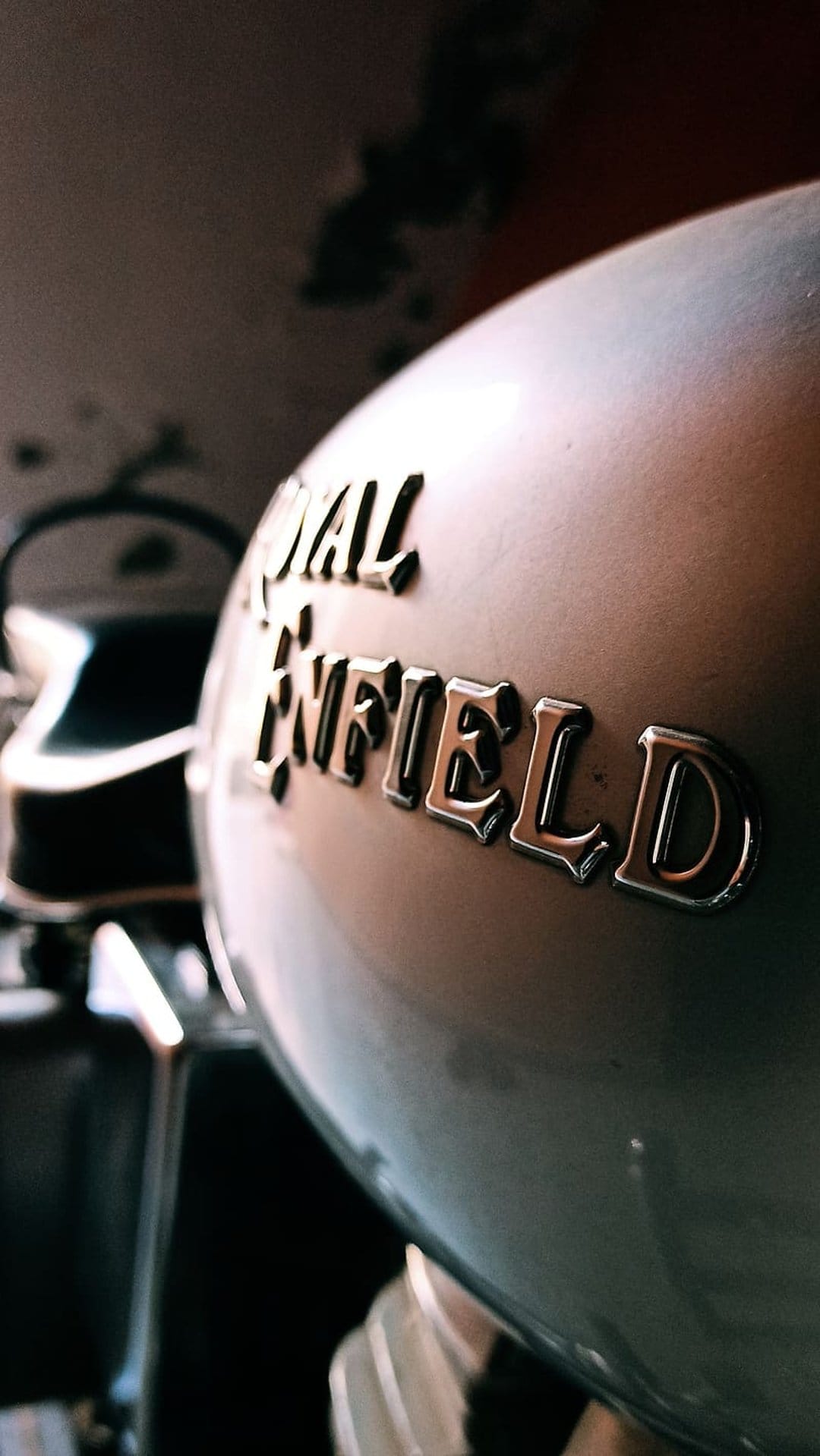 Royal Enfield chrome emblem on a metallic silver motorcycle tank with blurred mechanical parts in the background.
