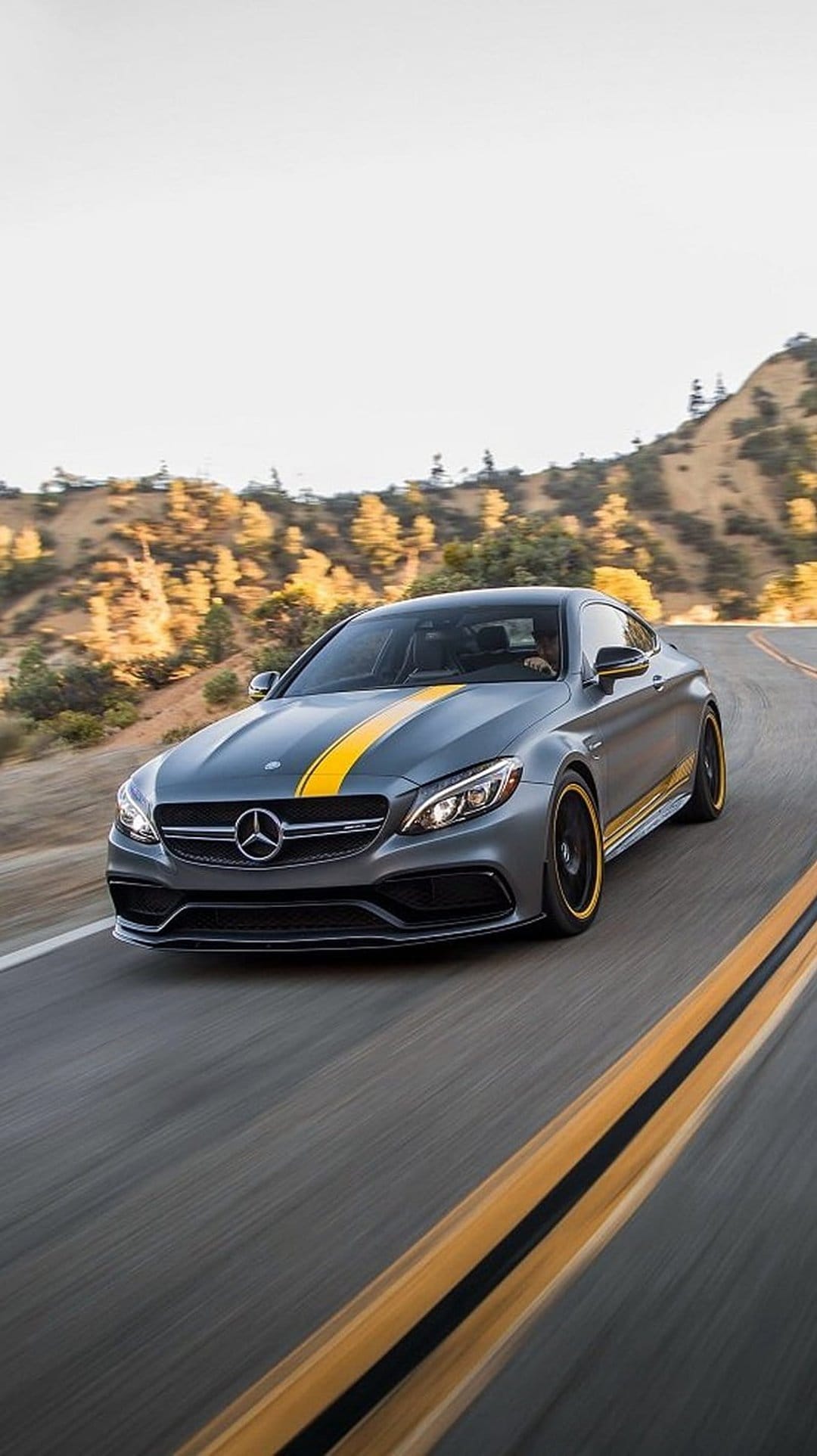 Mercedes-AMG C63 Coupe with yellow racing stripes drives on a winding mountain road under bright sunlight.