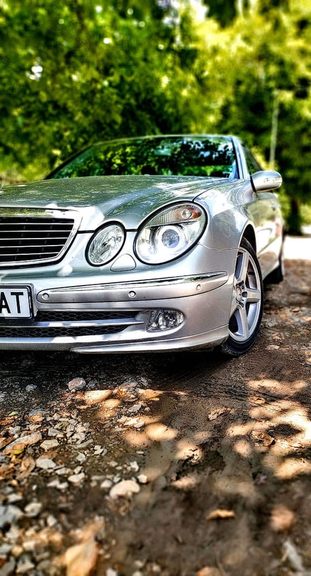 Silver Mercedes E-Class sedan parked on gravel under green trees with dappled sunlight hitting the front grille.
