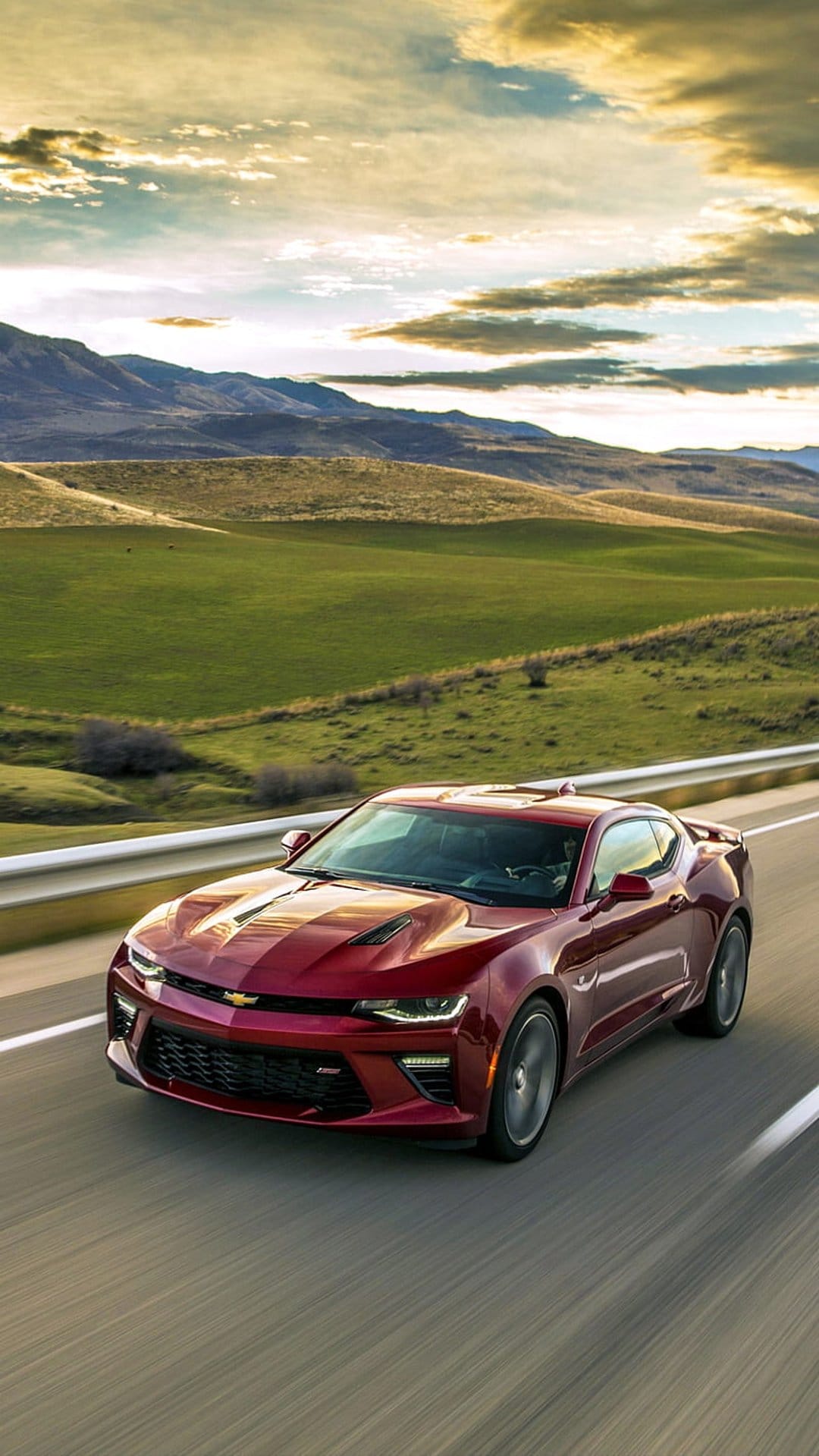 Red Chevrolet Camaro driving on a highway with rolling green hills and cloudy mountains in the background.