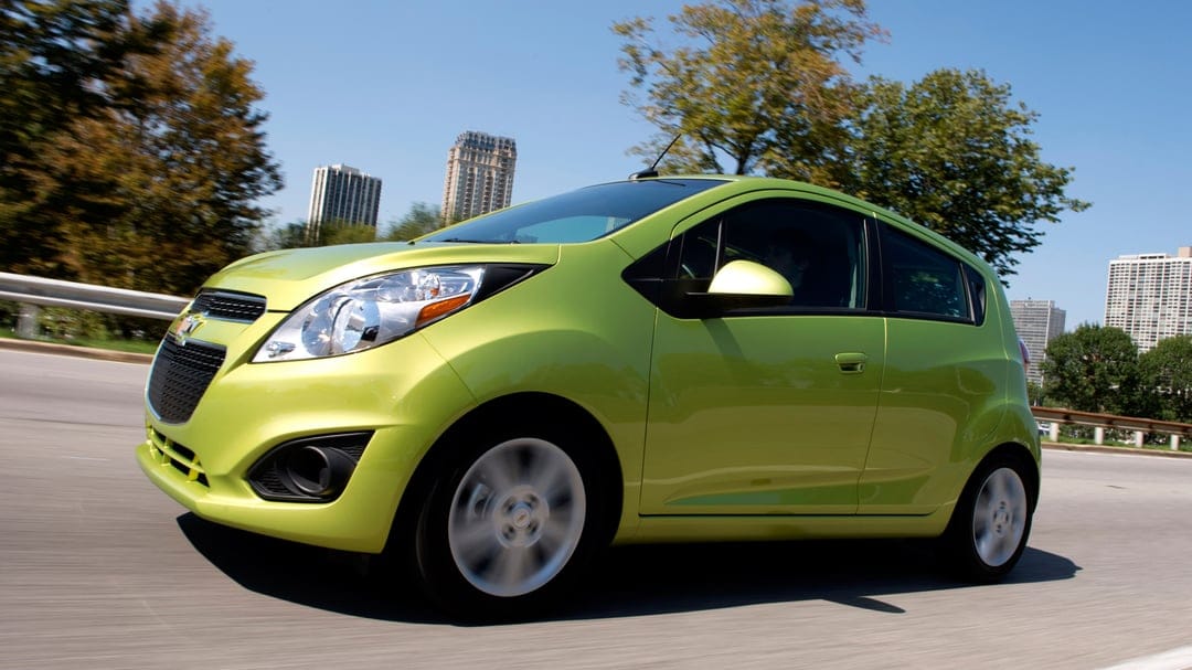 Lime green Chevrolet Spark hatchback driving on a city highway with skyscrapers and blue sky background.