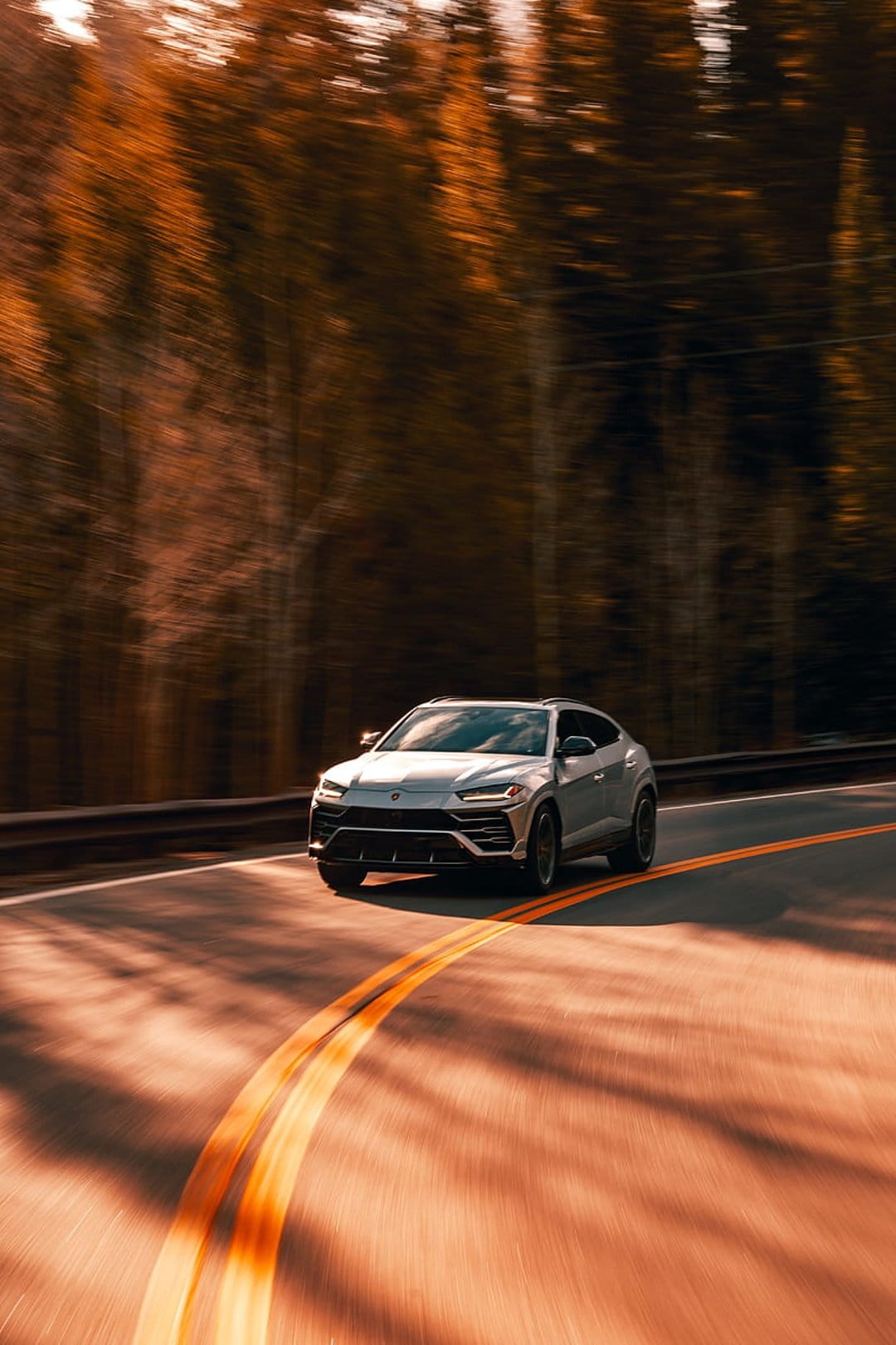 Silver Lamborghini Urus speeds on a winding road through an orange autumn forest with cinematic motion blur.