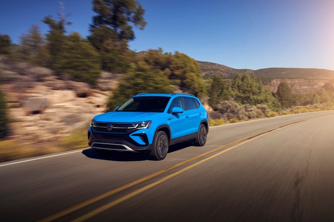 Blue modern SUV driving fast on a mountain highway with motion-blurred scenery under a clear blue sky.
