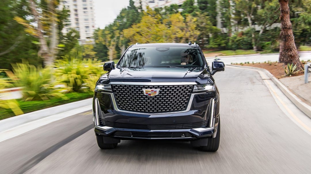 Dark blue Cadillac Escalade driving on a winding road with motion blur, green trees, and white buildings.