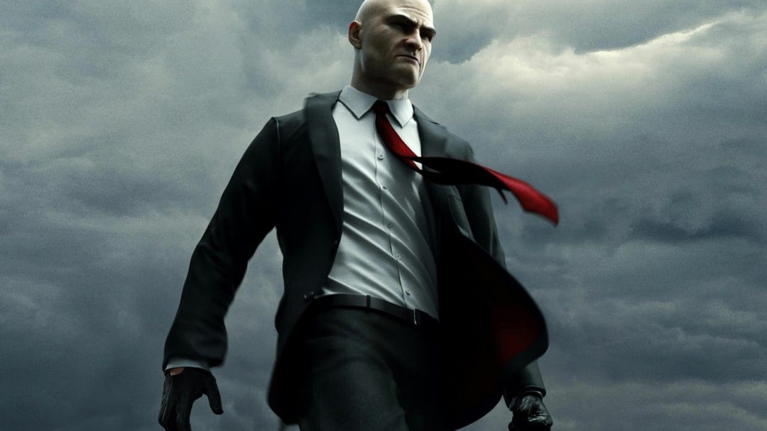 Agent 47 in a black suit with a red tie blowing in the wind against a dark, moody, overcast stormy sky.