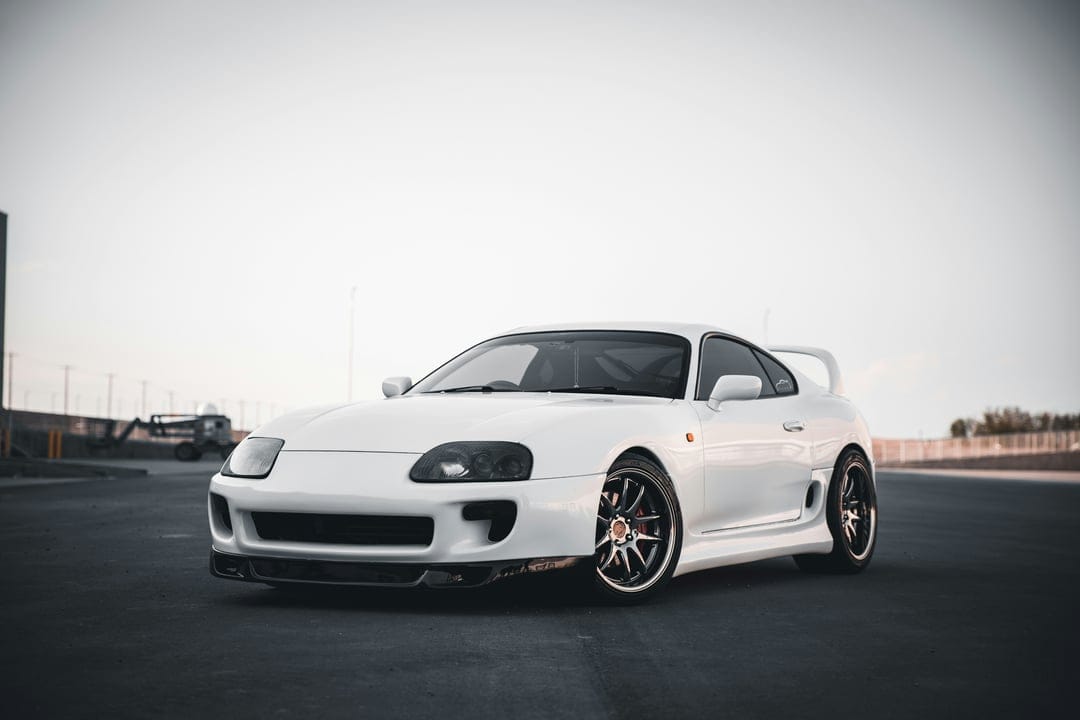 White Toyota Supra MK4 with black headlights and a front lip spoiler sits parked against a cloudy sky.