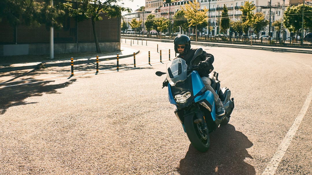 Blue scooter rider leans into a turn on a sunny city street with urban buildings and trees in the background.