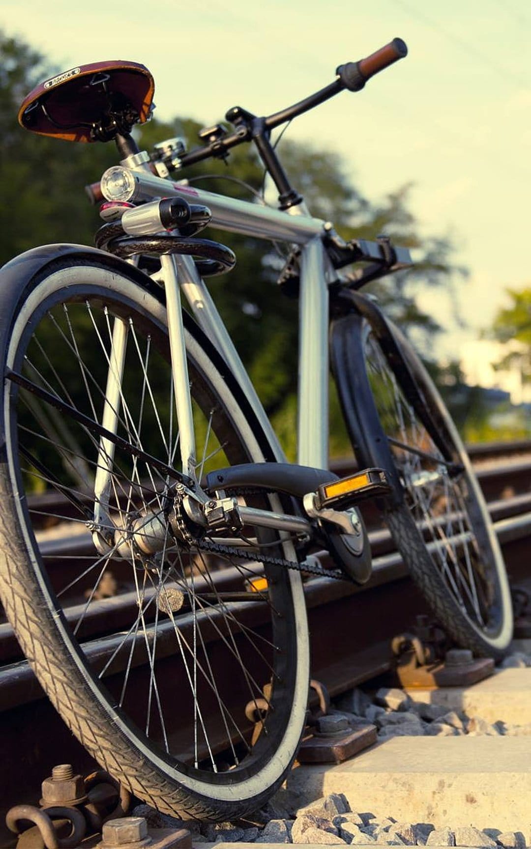 Silver bicycle with a brown leather seat resting on gravel-covered railroad tracks under warm sunlight.