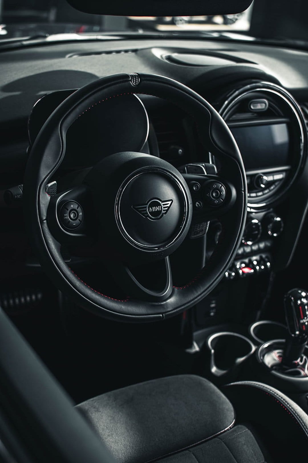 Mini Cooper steering wheel with red stitching and logo rests before a dark dashboard with a central display.