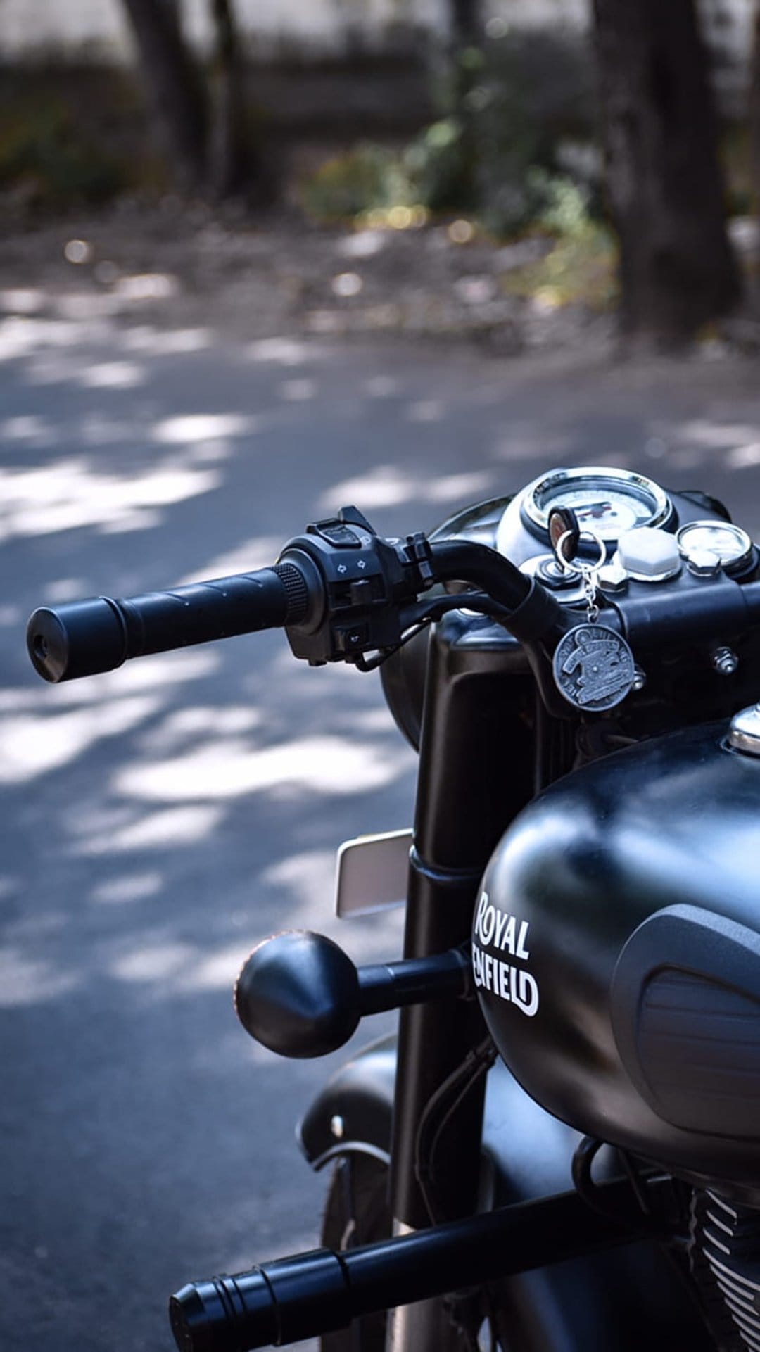 Matte black Royal Enfield fuel tank and handlebars with classic speedometer on a sunlit road for mobile screens.