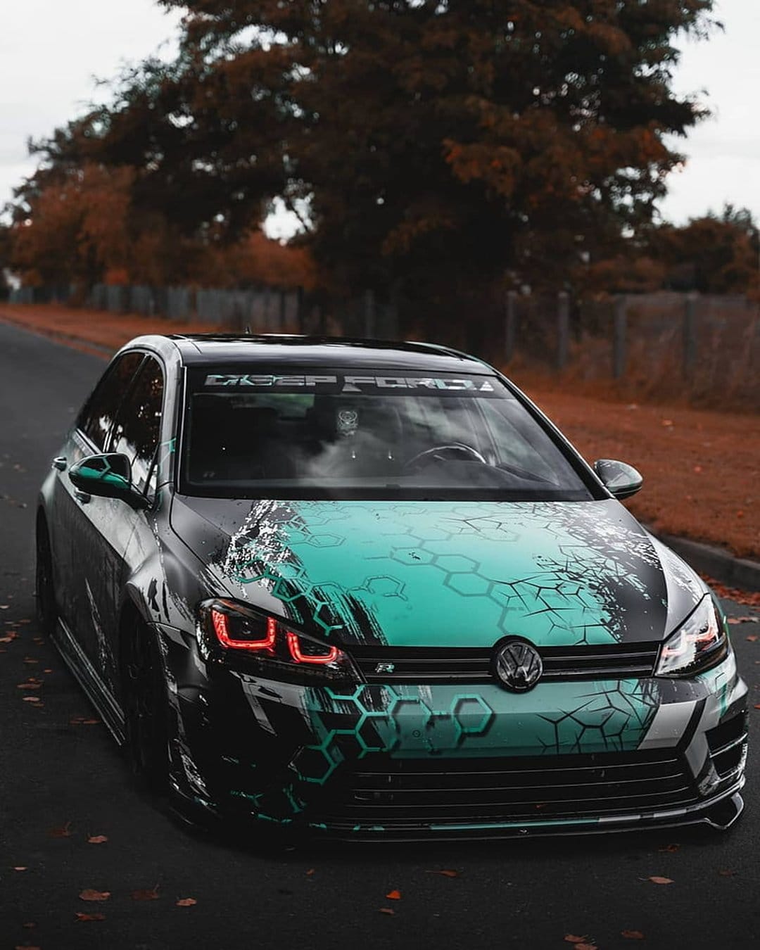 Volkswagen Golf R with teal hexagonal wrap and custom headlights parked on a leaf-covered autumn road.