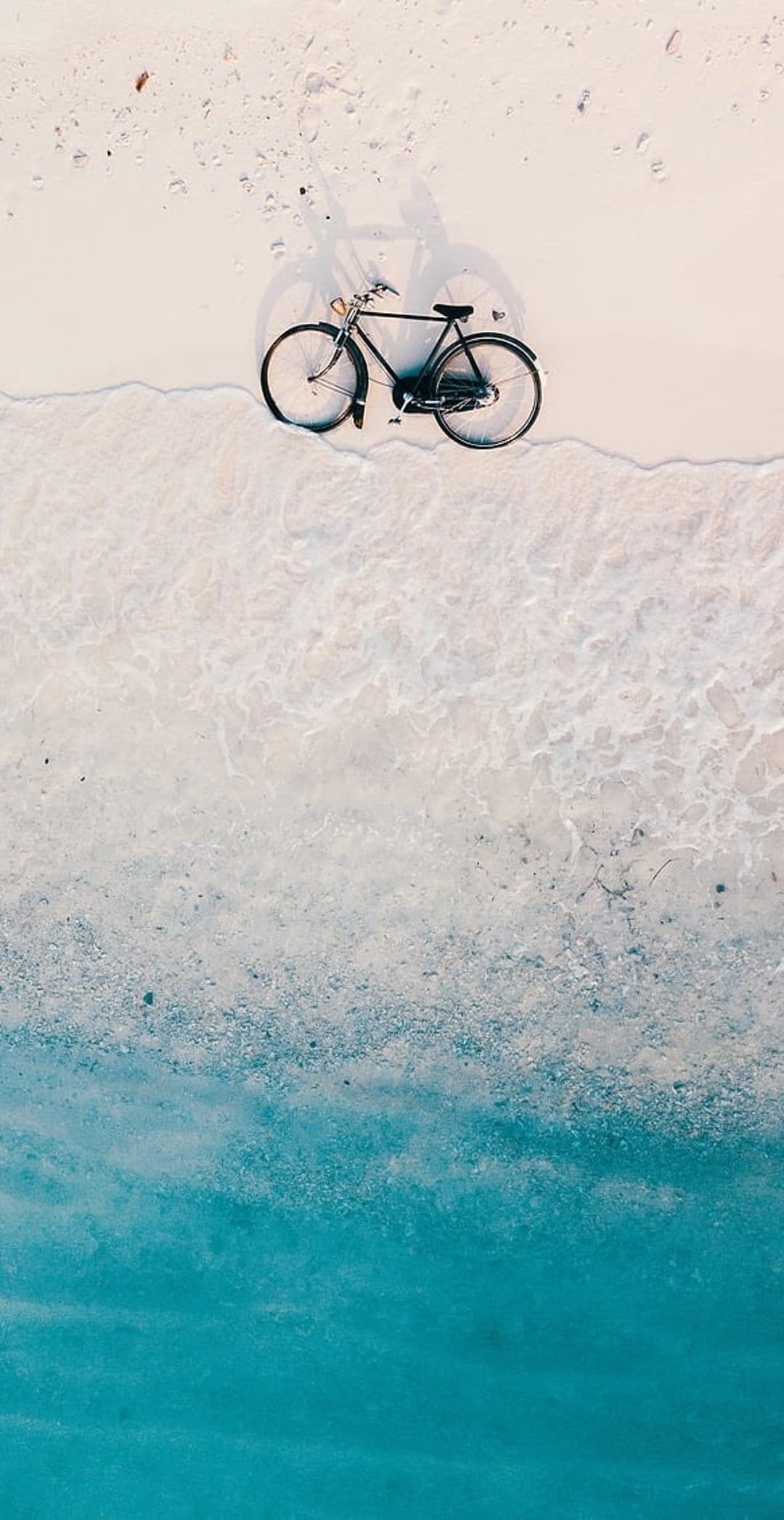 Black vintage bicycle parked on white sand next to turquoise ocean waves from a top-down aerial perspective.