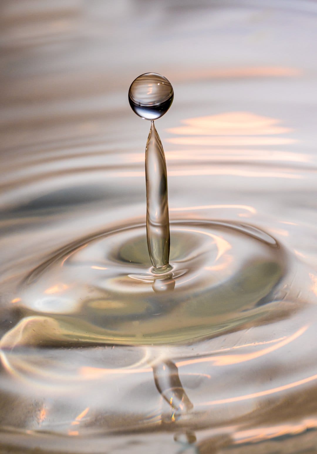 Spherical water drop balanced on a liquid column with expanding circular ripples under golden ambient light.