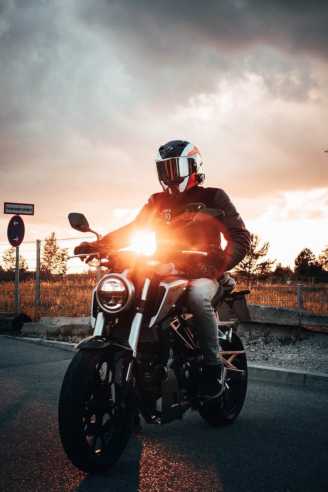 Motorcyclist on a road during golden hour with a bright lens flare and dramatic clouds in the background.