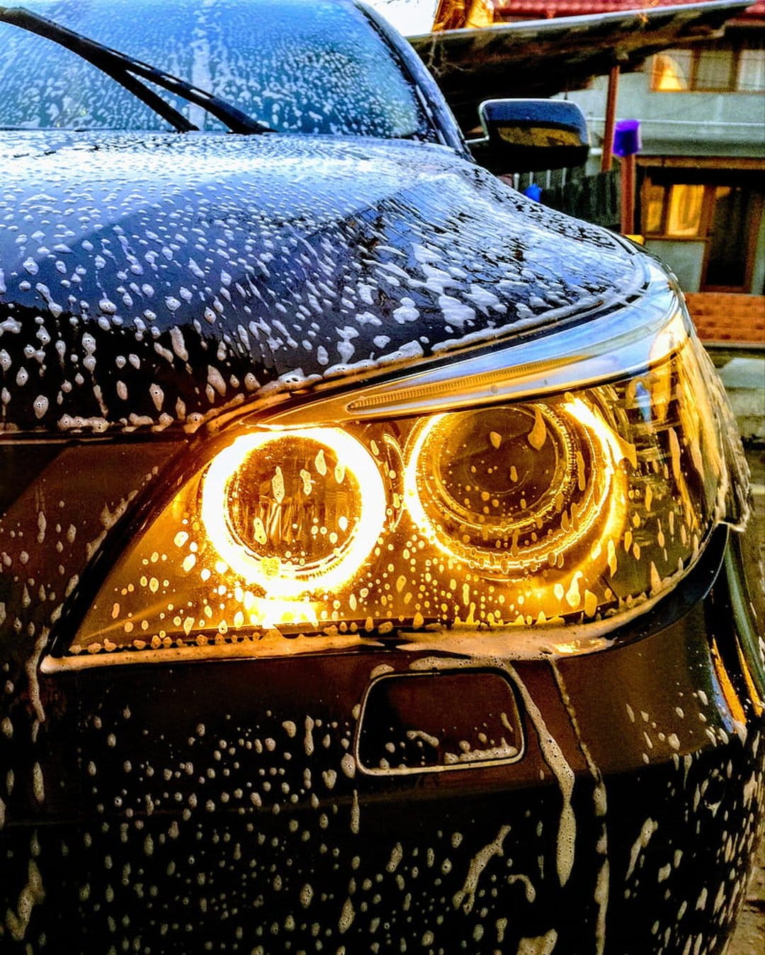 Orange car headlights glow intensely behind a thick layer of white soap bubbles on a dark, wet vehicle surface.