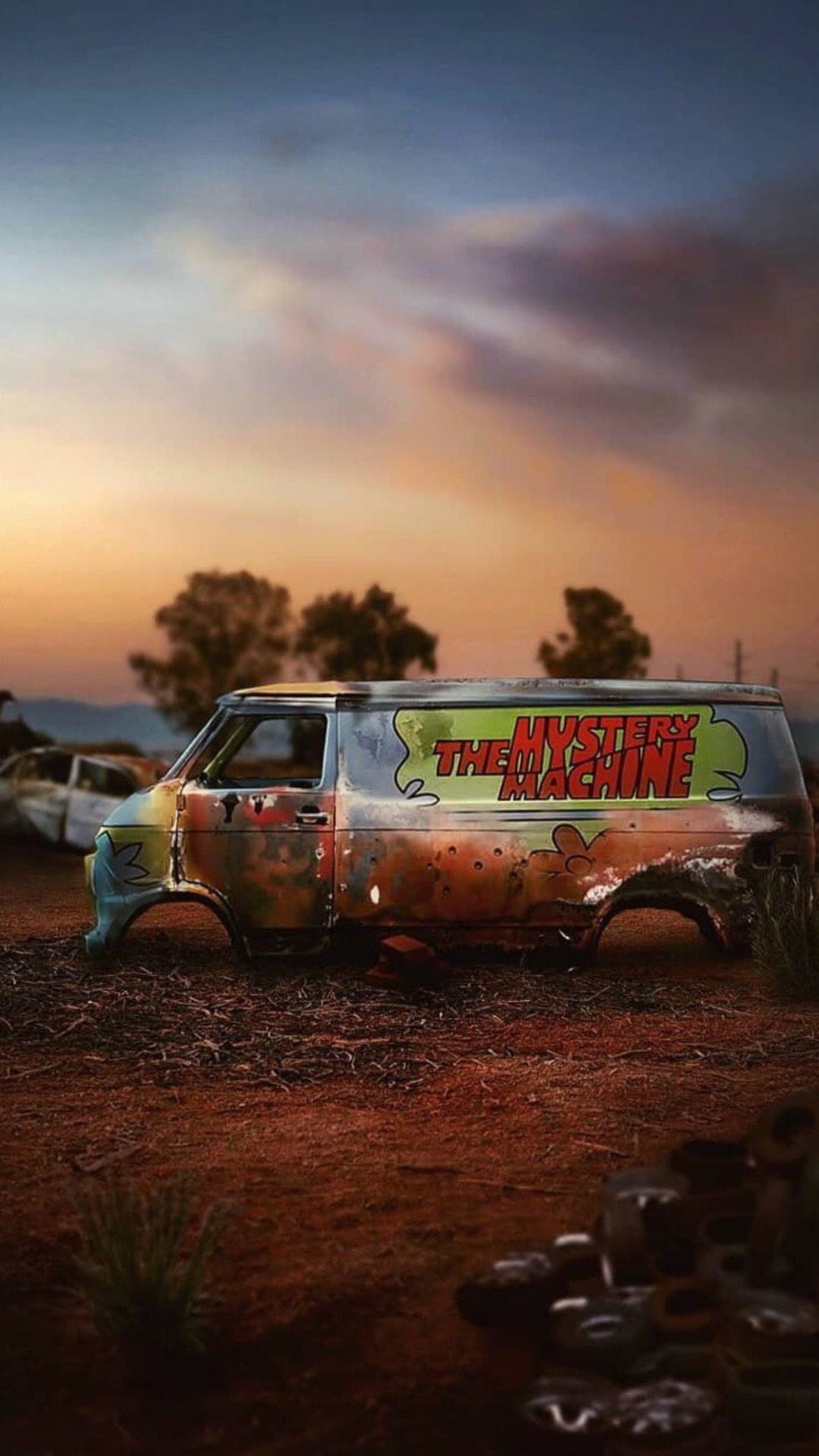 Mystery Machine van with rusted paint sits in a dusty desert under a vibrant orange and purple sunset sky.