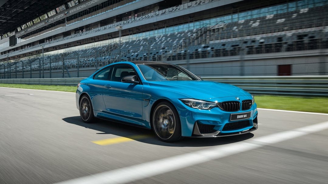 Blue BMW M4 sports car driving fast on a racetrack with motion blur and grandstands in the background.