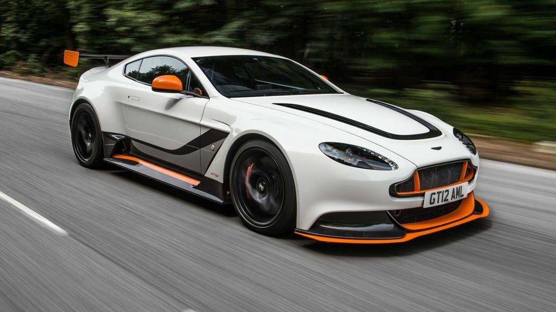 White Aston Martin GT12 with orange accents racing on a road with motion blurred trees and wheels.