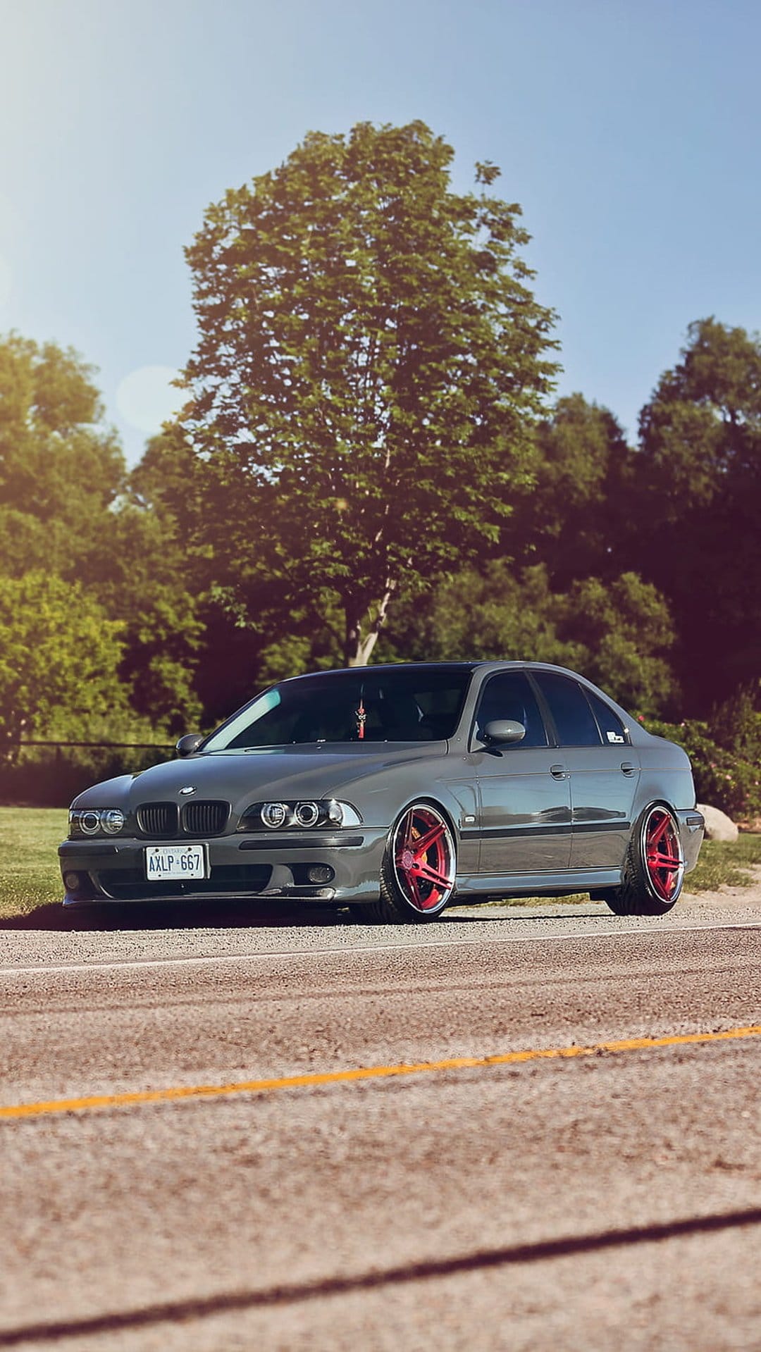 Gray BMW E39 M5 sedan with red aftermarket wheels on an asphalt road surrounded by green summer trees.