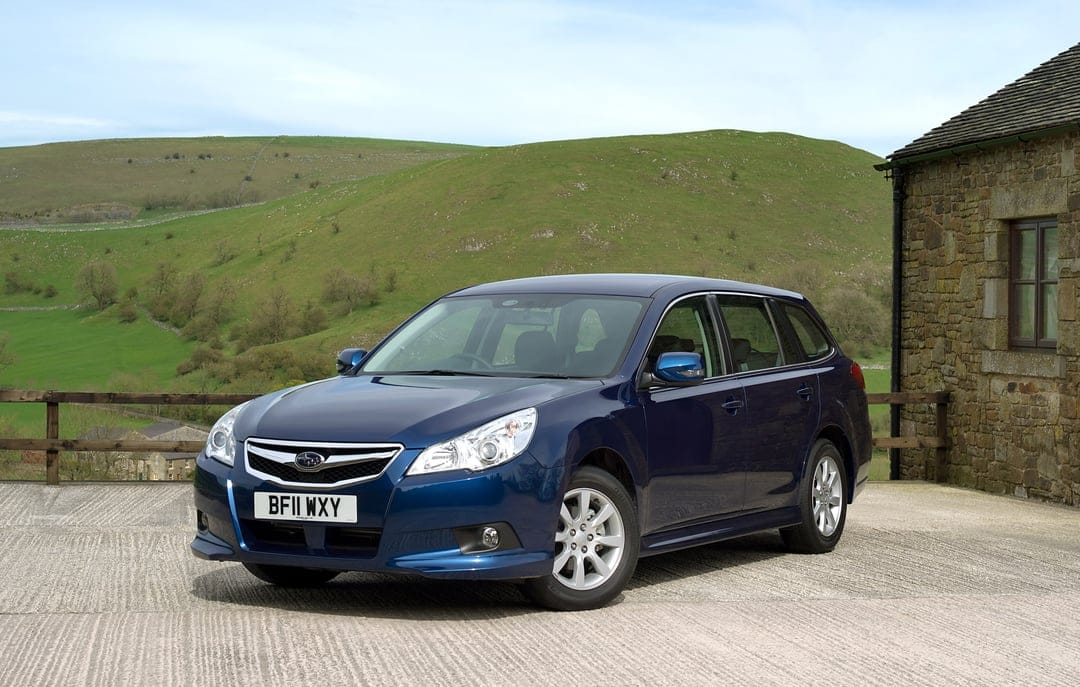 Dark blue Subaru Legacy wagon parked on a concrete lot against rolling green hills and a stone building.