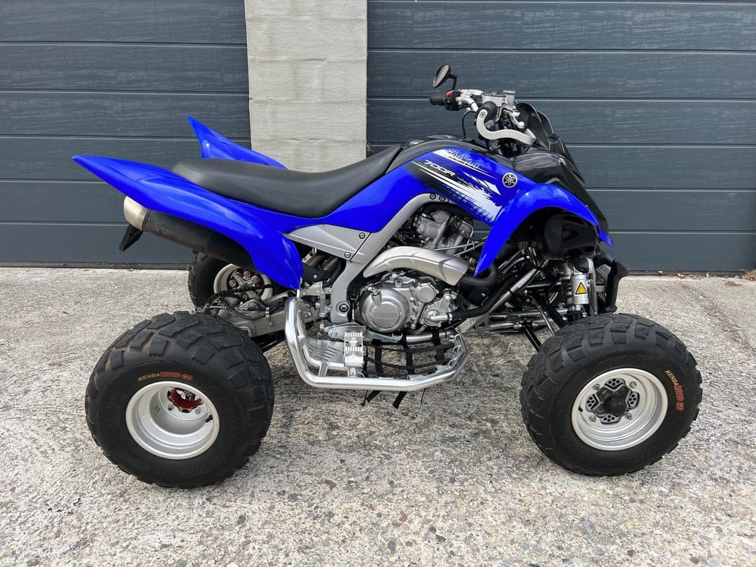 Blue Yamaha Raptor 700R sport ATV side profile parked on a concrete driveway in front of a gray garage door.